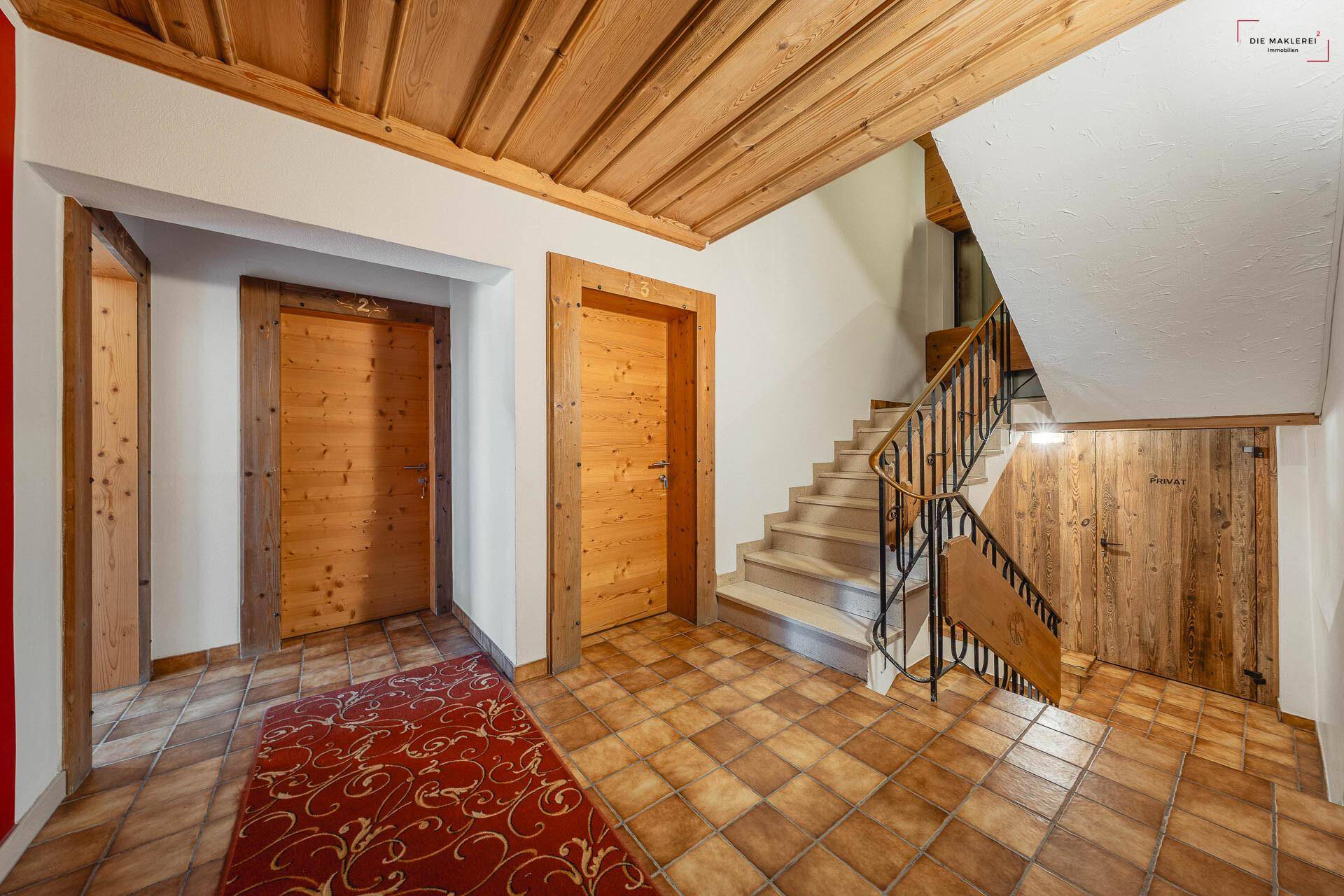 Rustikaler Flur mit Holzdecke, Fliesenboden und einer Treppe mit Holzgeländer.