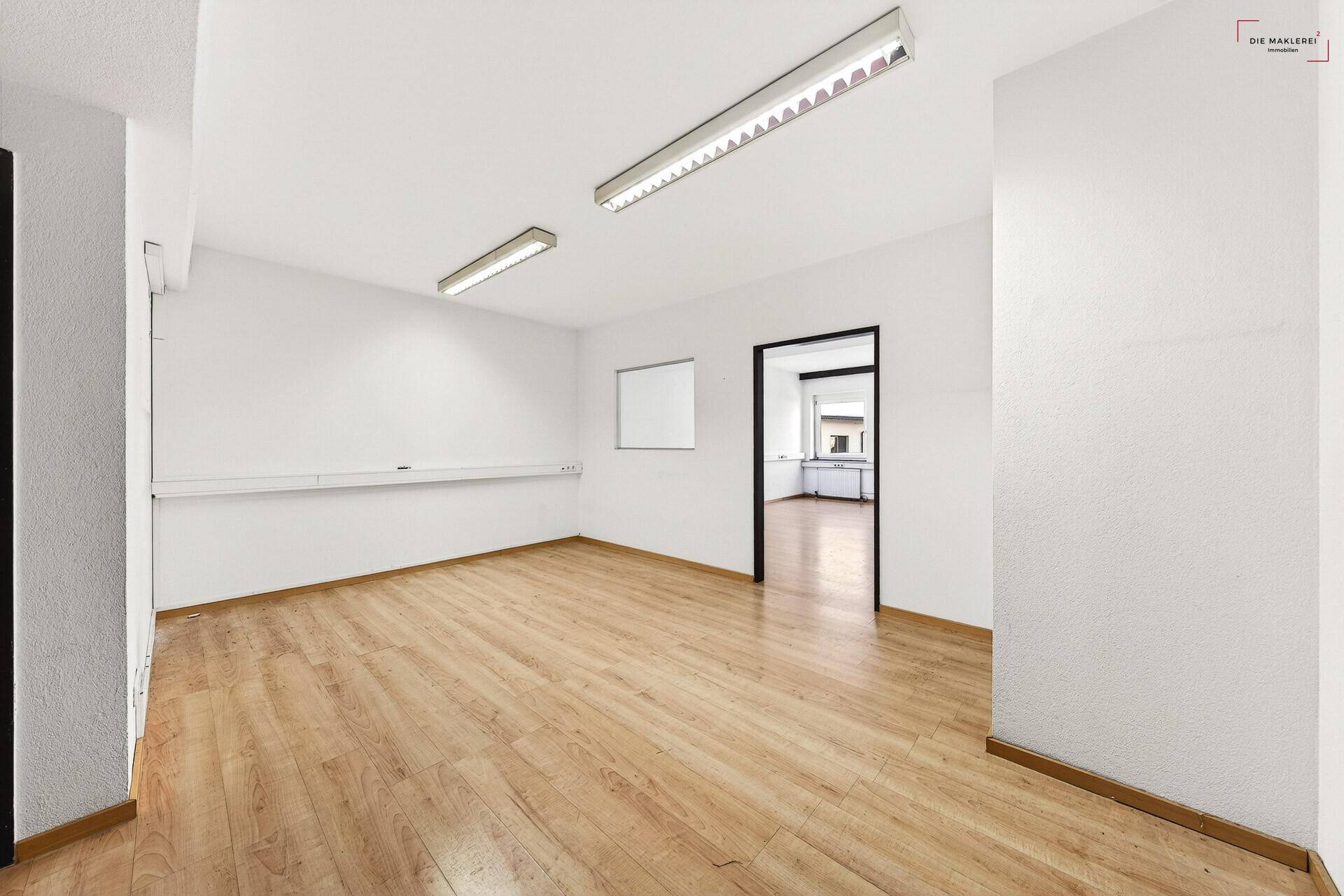 Heller Büroraum mit Laminatboden, weißen Wänden und einem offenen Durchgang zu einem weiteren Bereich.