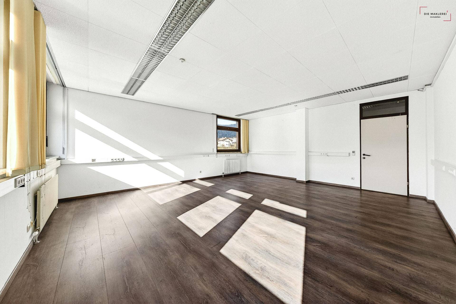Geräumiger Büroraum mit dunklem Laminatboden und hellen Wänden, durchflutet von Sonnenlicht.