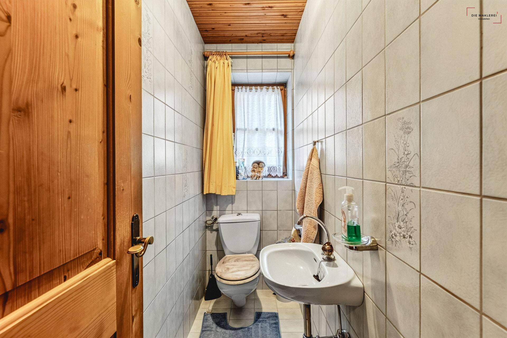 Kleines Gäste-WC mit Fliesen, Waschbecken, Toilette und Fenster mit Vorhang.
