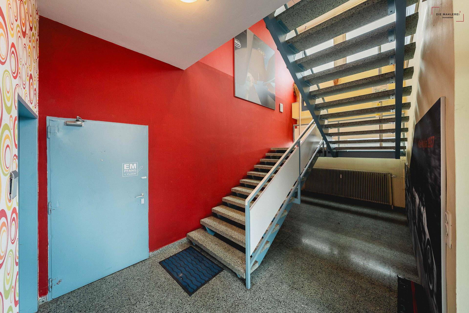 Treppenhaus mit roter Wand, robuster Metalltreppe und Terrazzoboden, der zu den oberen Etagen führt.