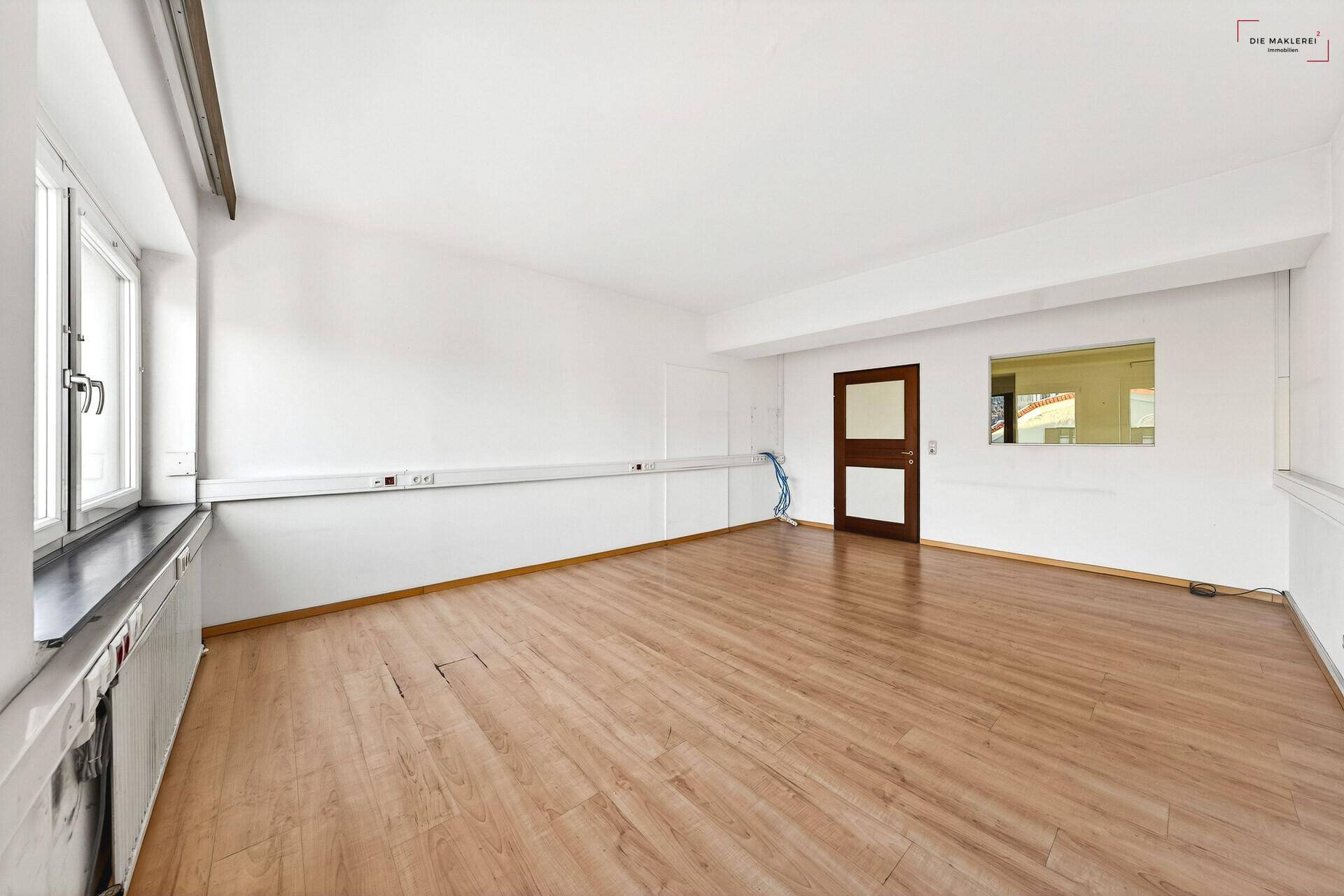 Leerer Büroraum mit Laminatboden, weißen Wänden und einer langen Ablagefläche unter den Fenstern.