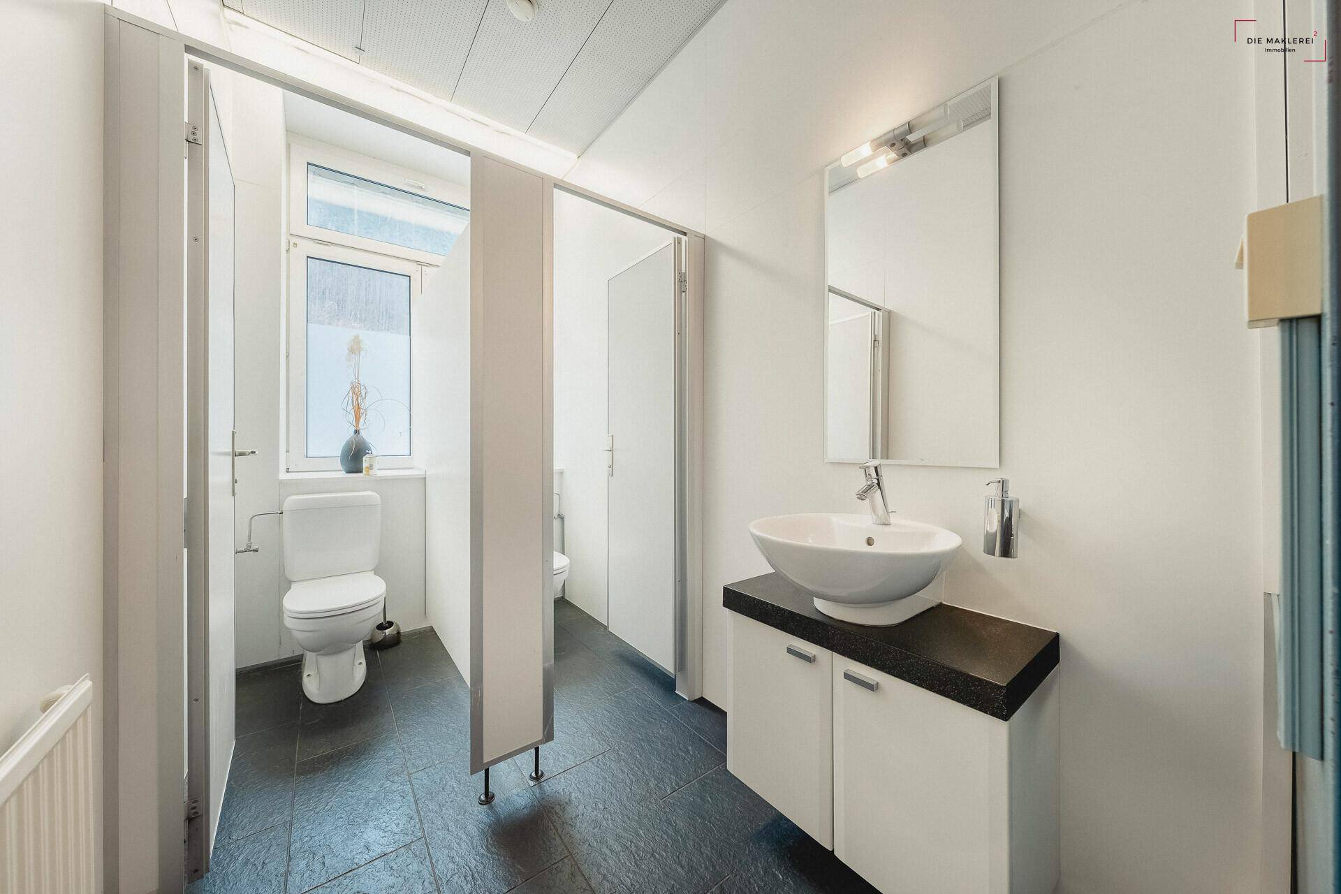 Moderner Waschraum mit mehreren Toilettenkabinen, Waschbecken und dunklen Fliesen, gut beleuchtet.