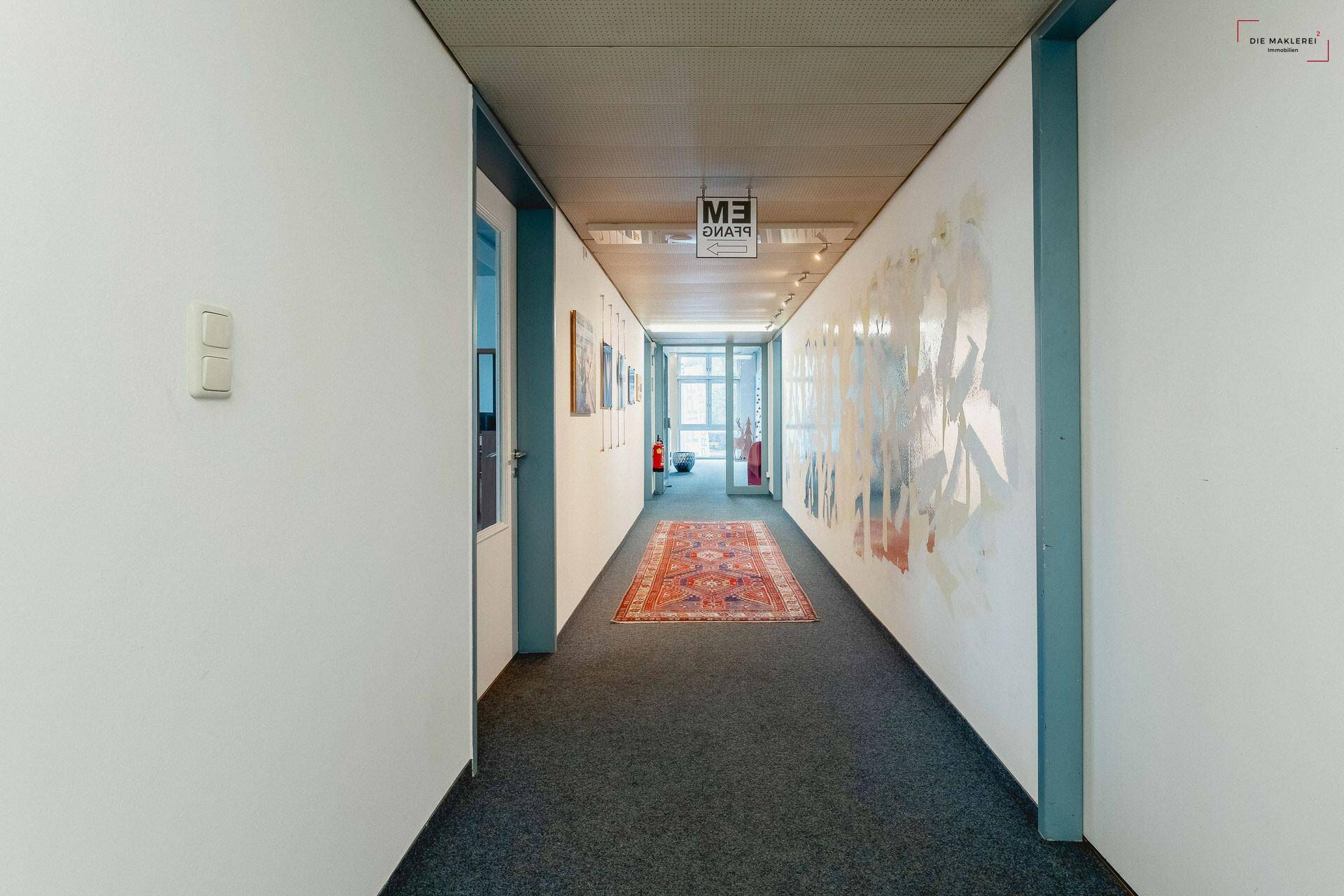 Langer Flur mit Teppichboden, Kunst an der Wand und einem Fenster am Ende, das Tageslicht hereinlässt.