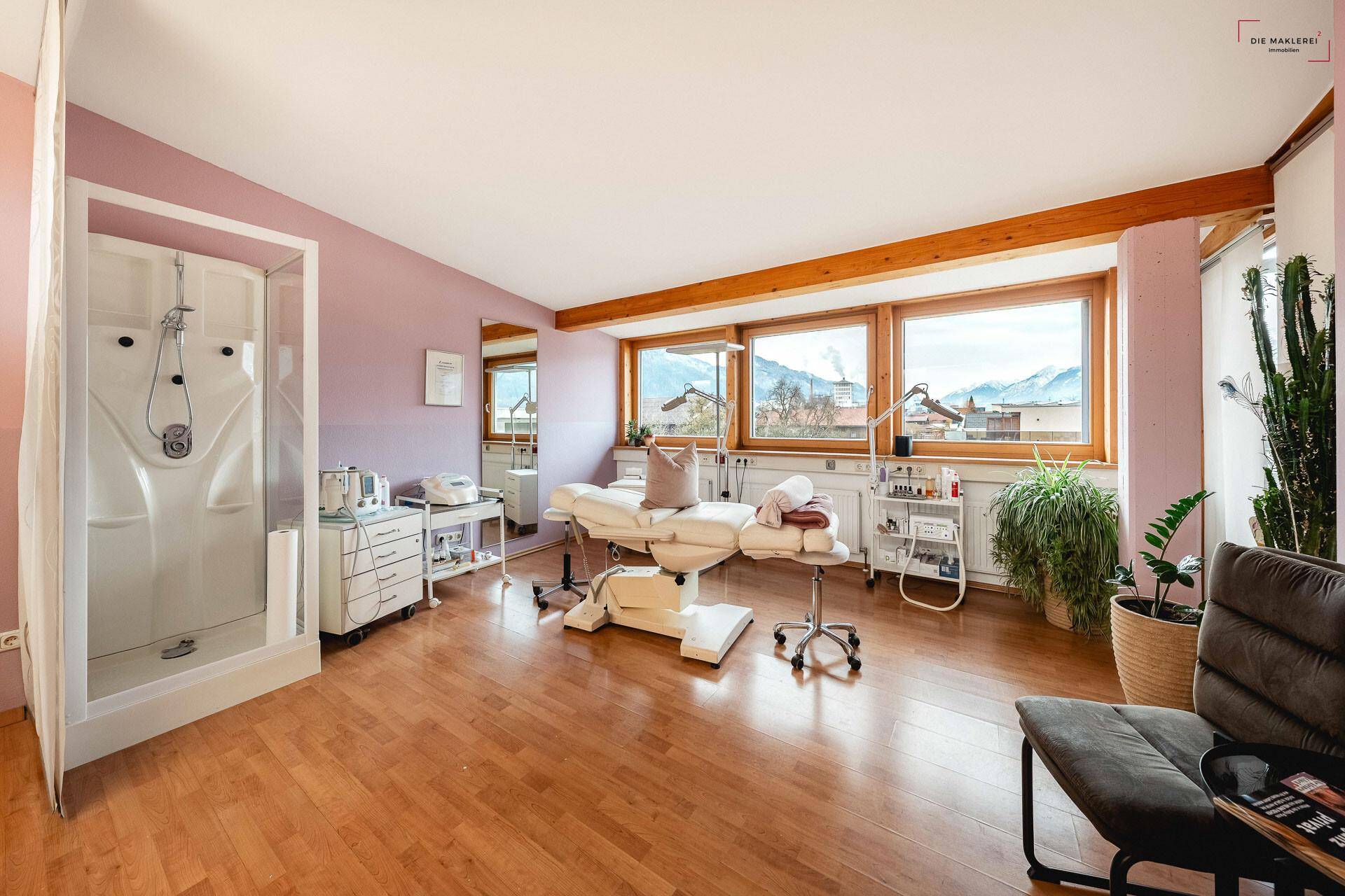 Heller Behandlungsraum mit Holzboden, Duschkabine und Panoramablick auf die Berge durch große Fenster.