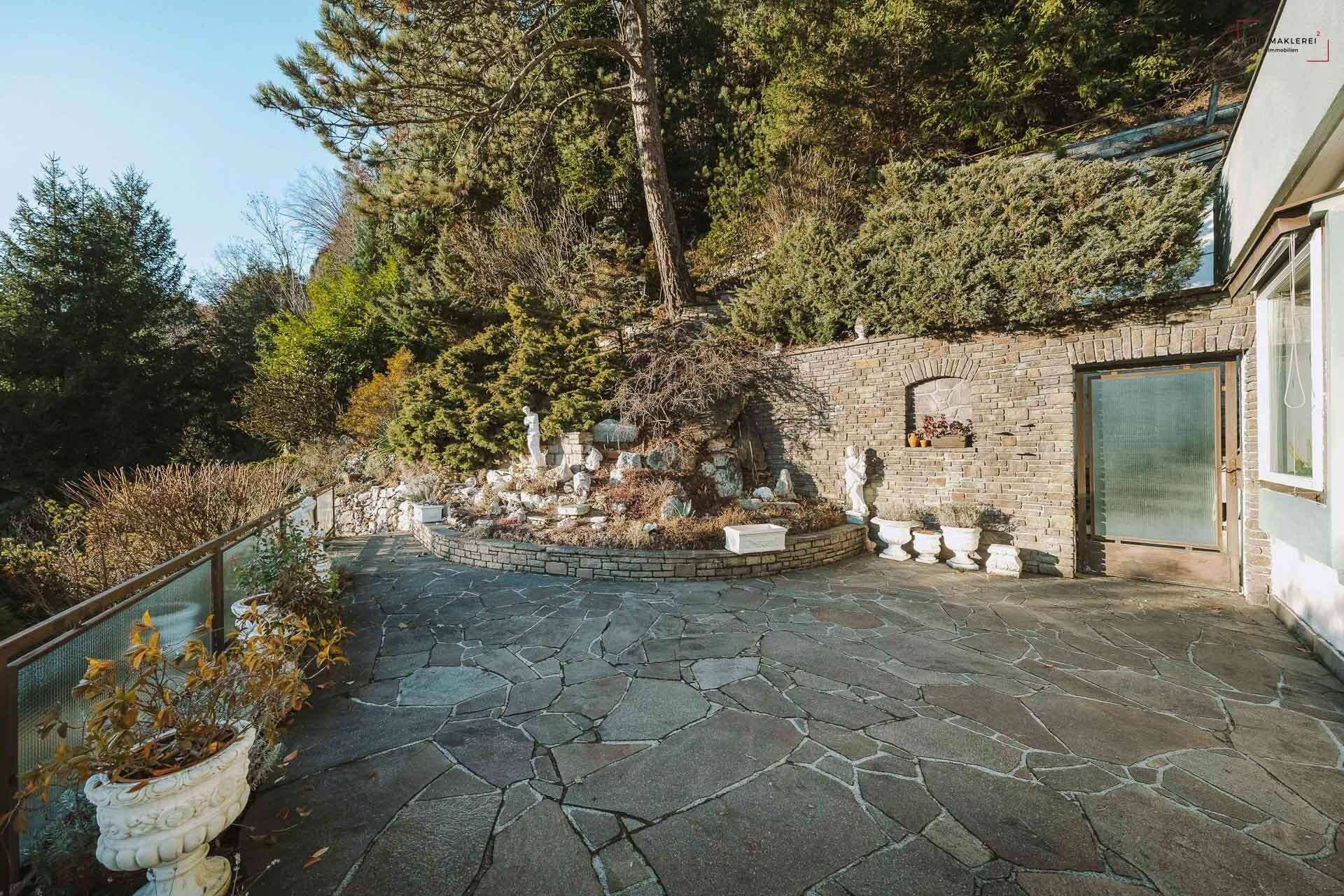 Terrassierter Gartenbereich mit Natursteinplatten, einer Steinmauer und dekorativen Statuen.