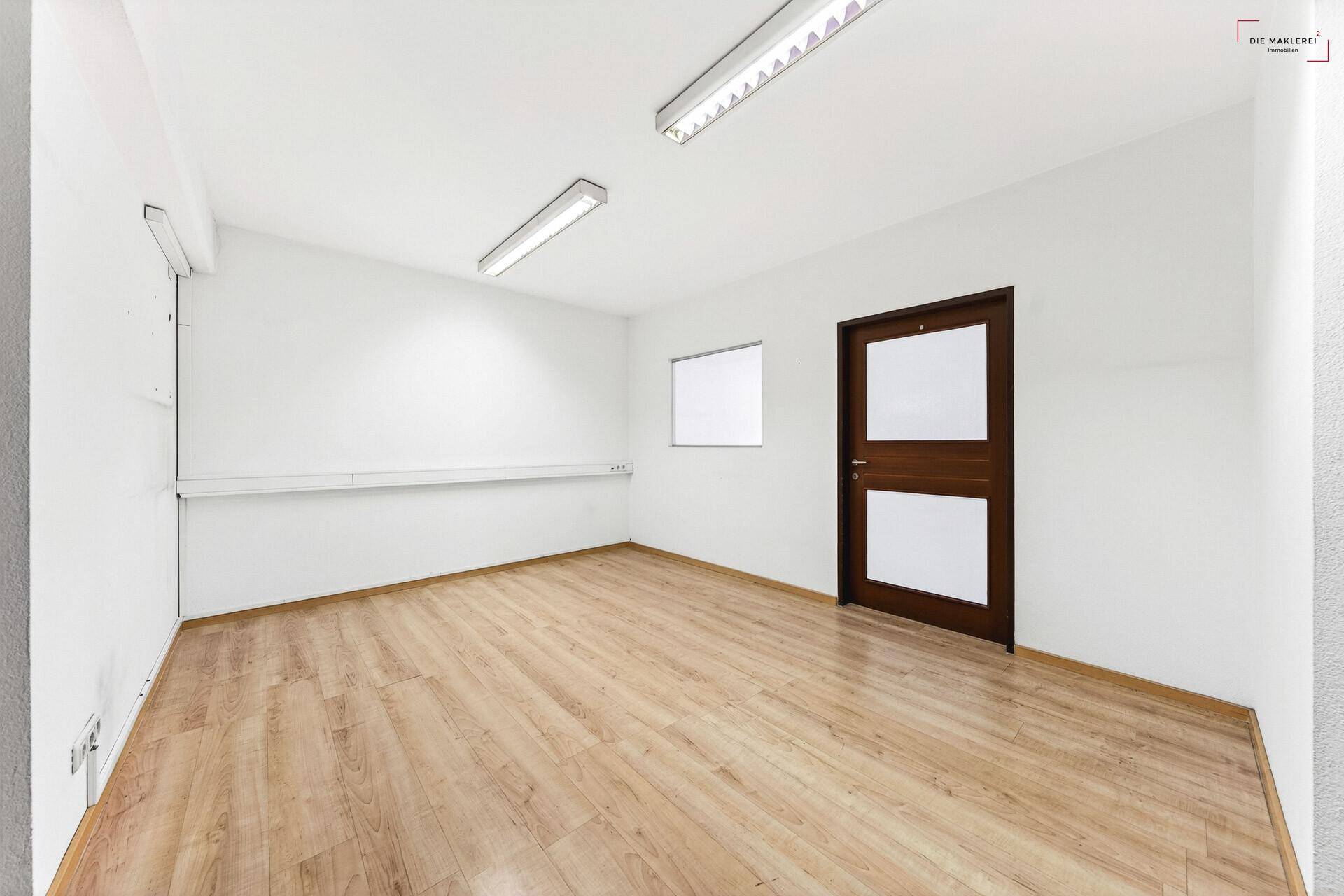 Geräumiger Büroraum mit Laminatboden, weißen Wänden und einer modernen Holztür mit Glaseinsatz.
