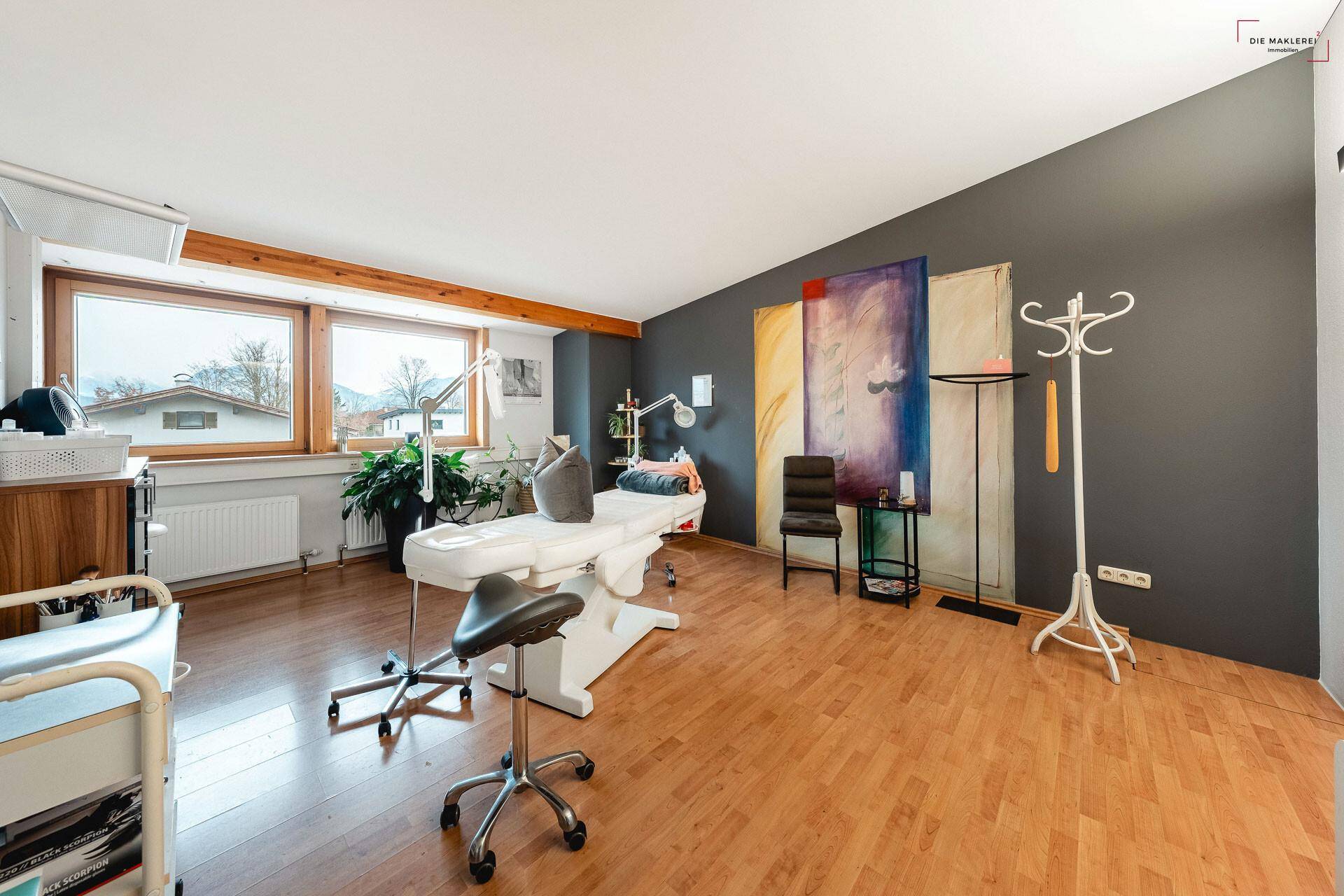 Geräumiger Behandlungsraum mit Holzboden, großen Fenstern und dunkler Akzentwand, ausgestattet für Therapien.