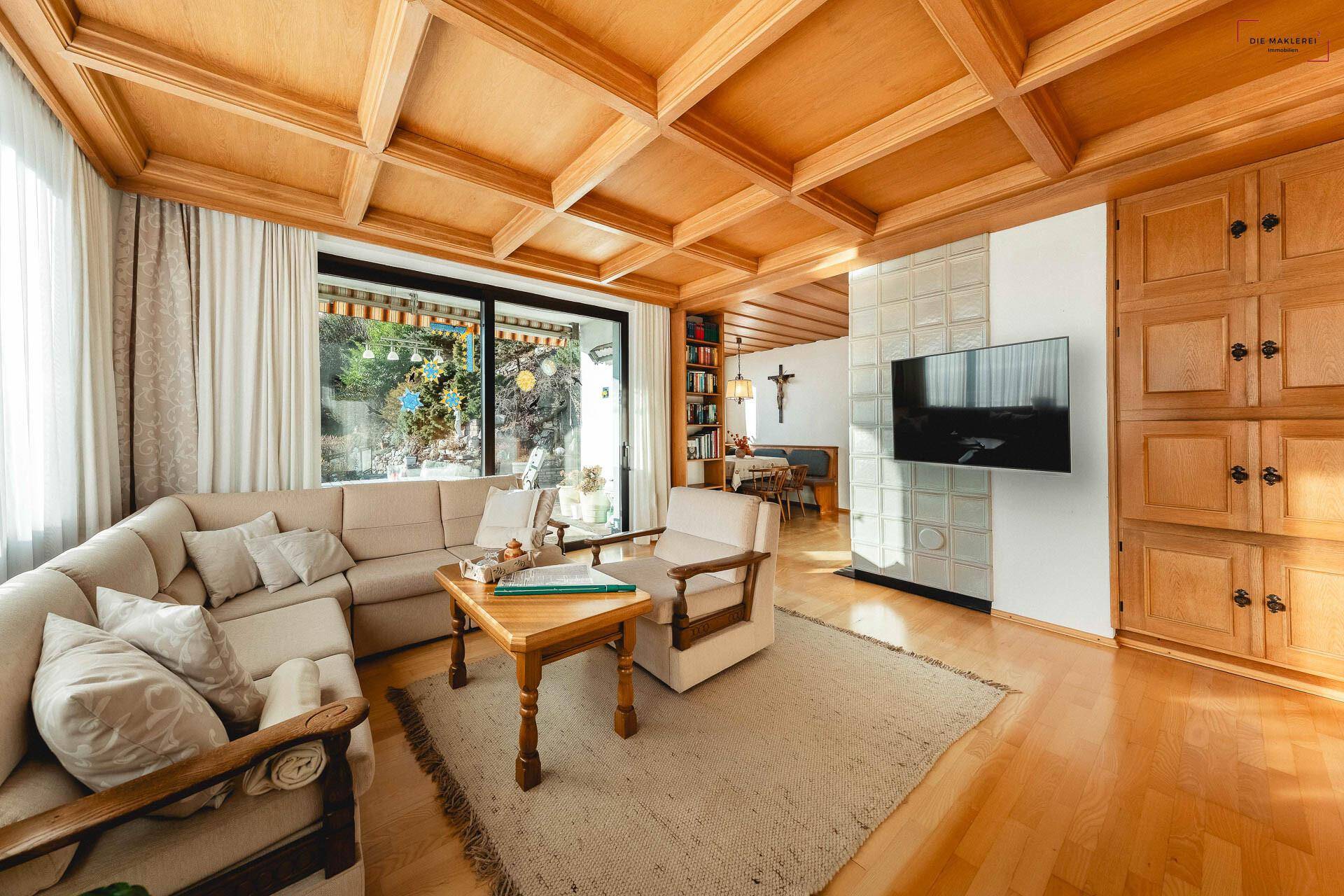 Gemütliches Wohnzimmer mit Holzkassettendecke, großen Fenstern und bequemen Sitzmöbeln.