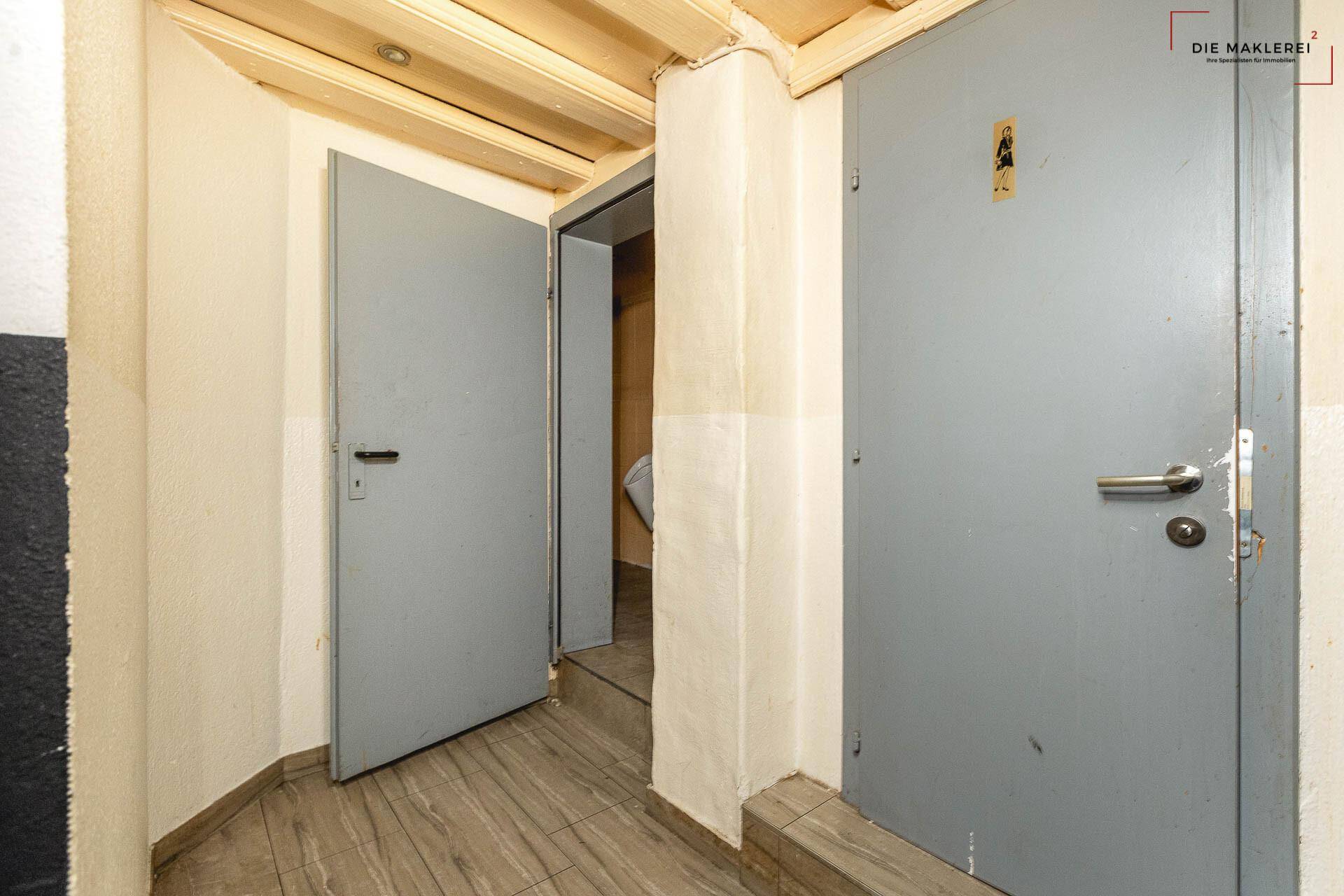 Eingangsbereich zu den Toiletten mit zwei grauen Türen und Holzbalkendecke.