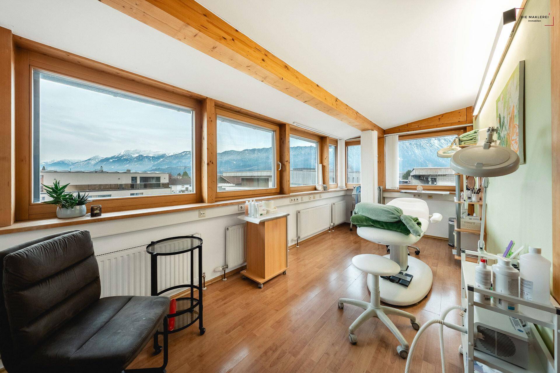Heller Behandlungsraum mit Holzboden, großen Fenstern und weitem Blick auf die Berglandschaft.