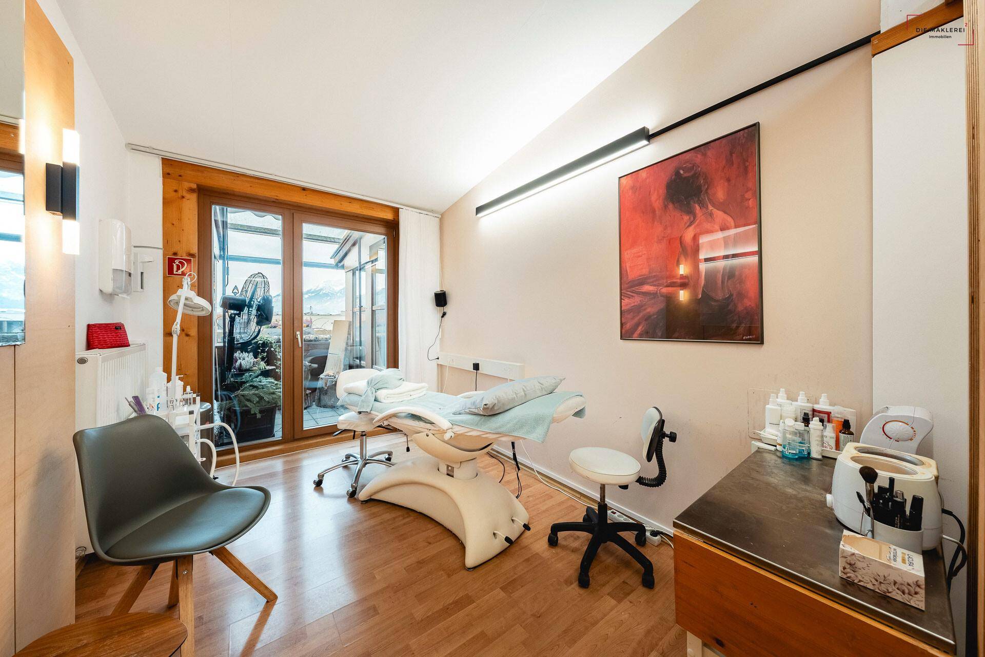 Gemütlicher Behandlungsraum mit Holzboden, Kunstwerk und Zugang zu einem überdachten Balkon mit Bergblick.