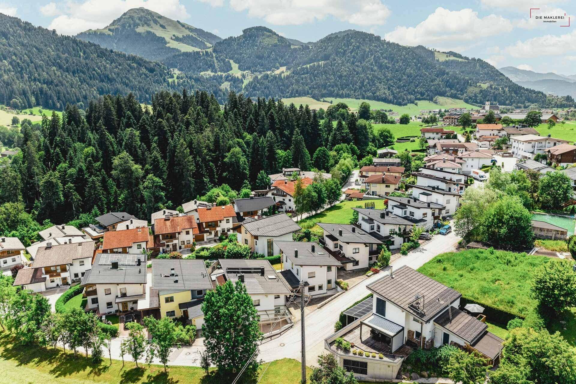 Luftaufnahme einer Wohnsiedlung mit Häusern und grüner Berglandschaft im Hintergrund.
