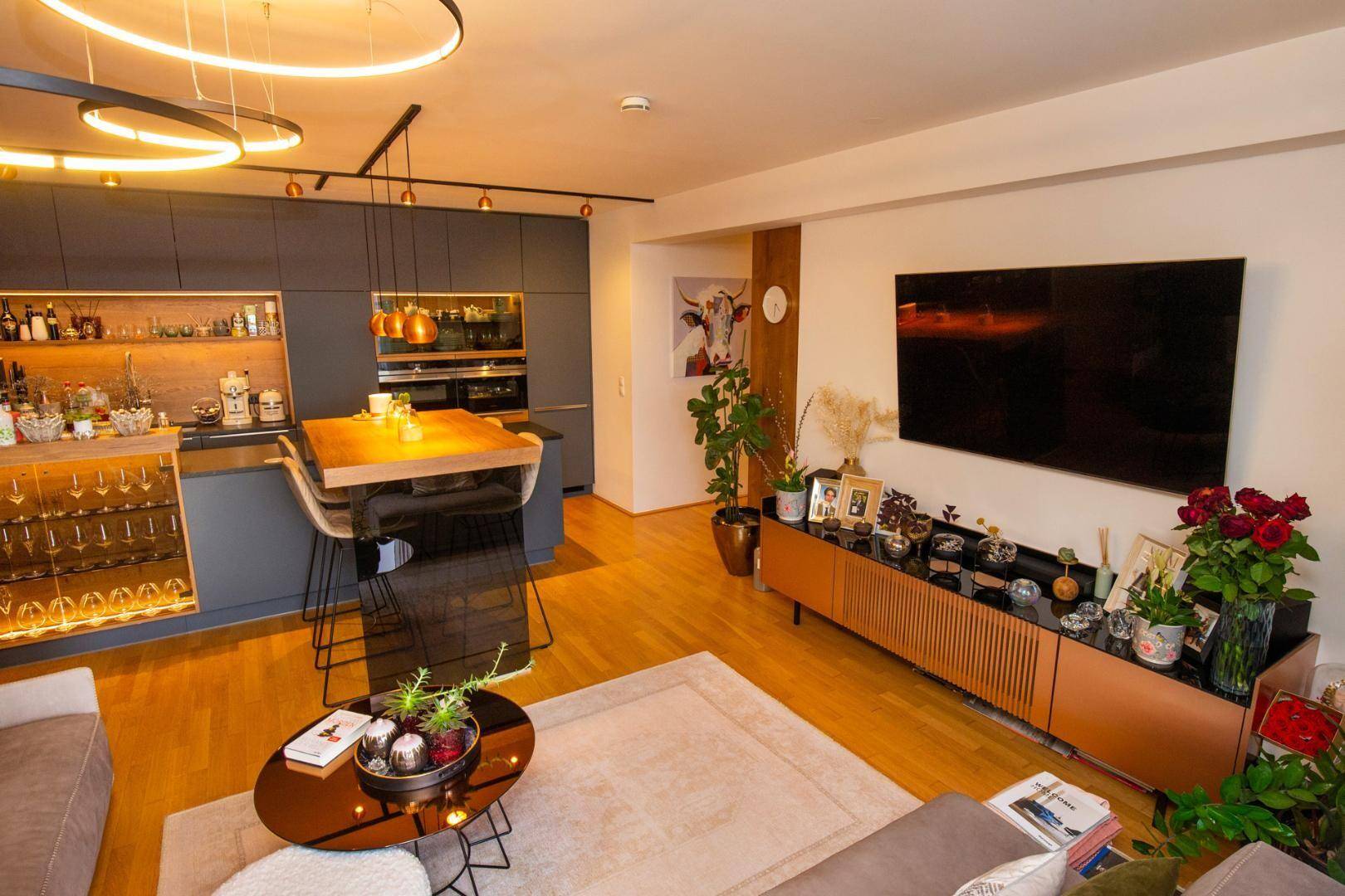 Offener Wohnbereich mit Fernseher, Holzboden und moderner Küche im Hintergrund.
