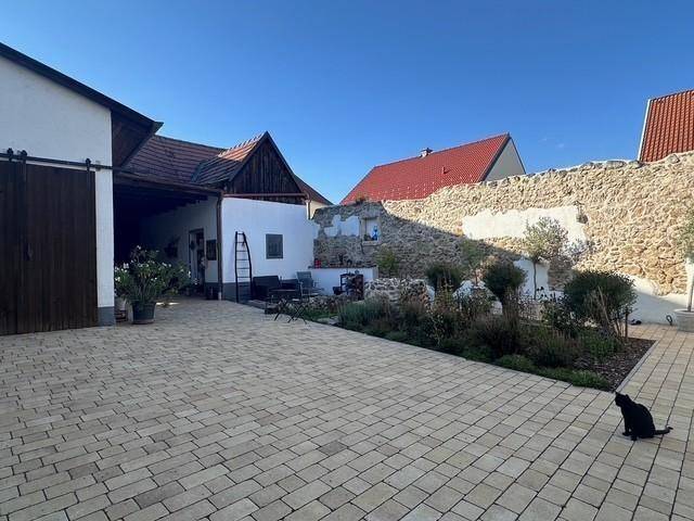 Weiter Blick über den gepflasterten Innenhof mit Steinmauer, Bepflanzung und einem schwarzen Kater.