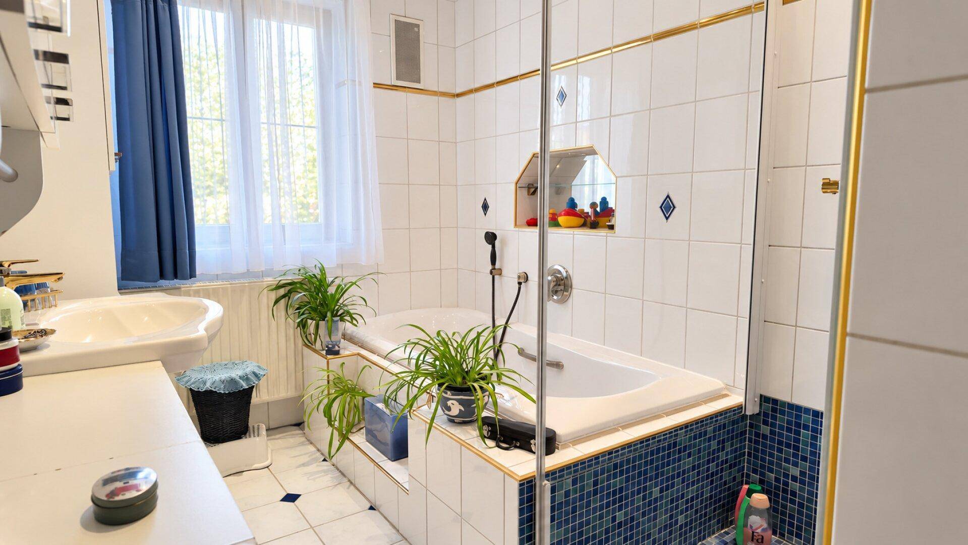 Helles Badezimmer mit Badewanne, Dusche, Fenster und weißen Fliesen mit goldenen Akzenten.