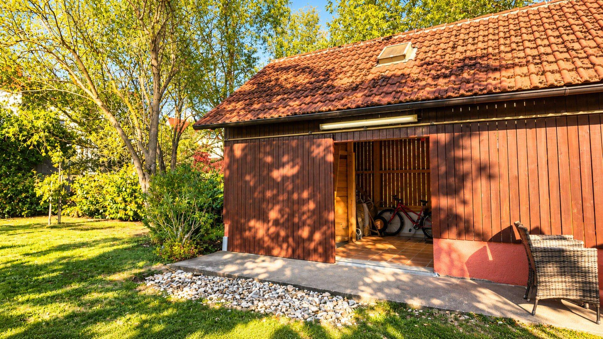 Geräumiger Holzschuppen im Garten, teilweise geöffnet, mit Fahrrädern und Gartenmöbeln davor.