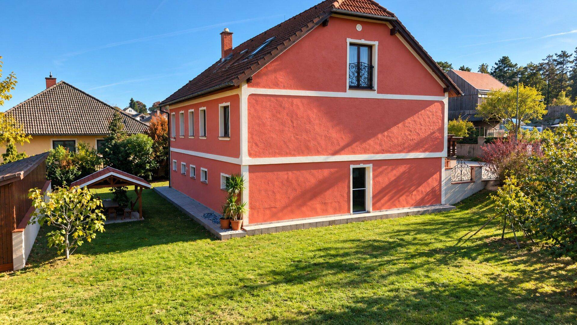 Seitenansicht des roten Wohnhauses mit gepflegtem Rasen und sonnigem Gartenbereich unter blauem Himmel.