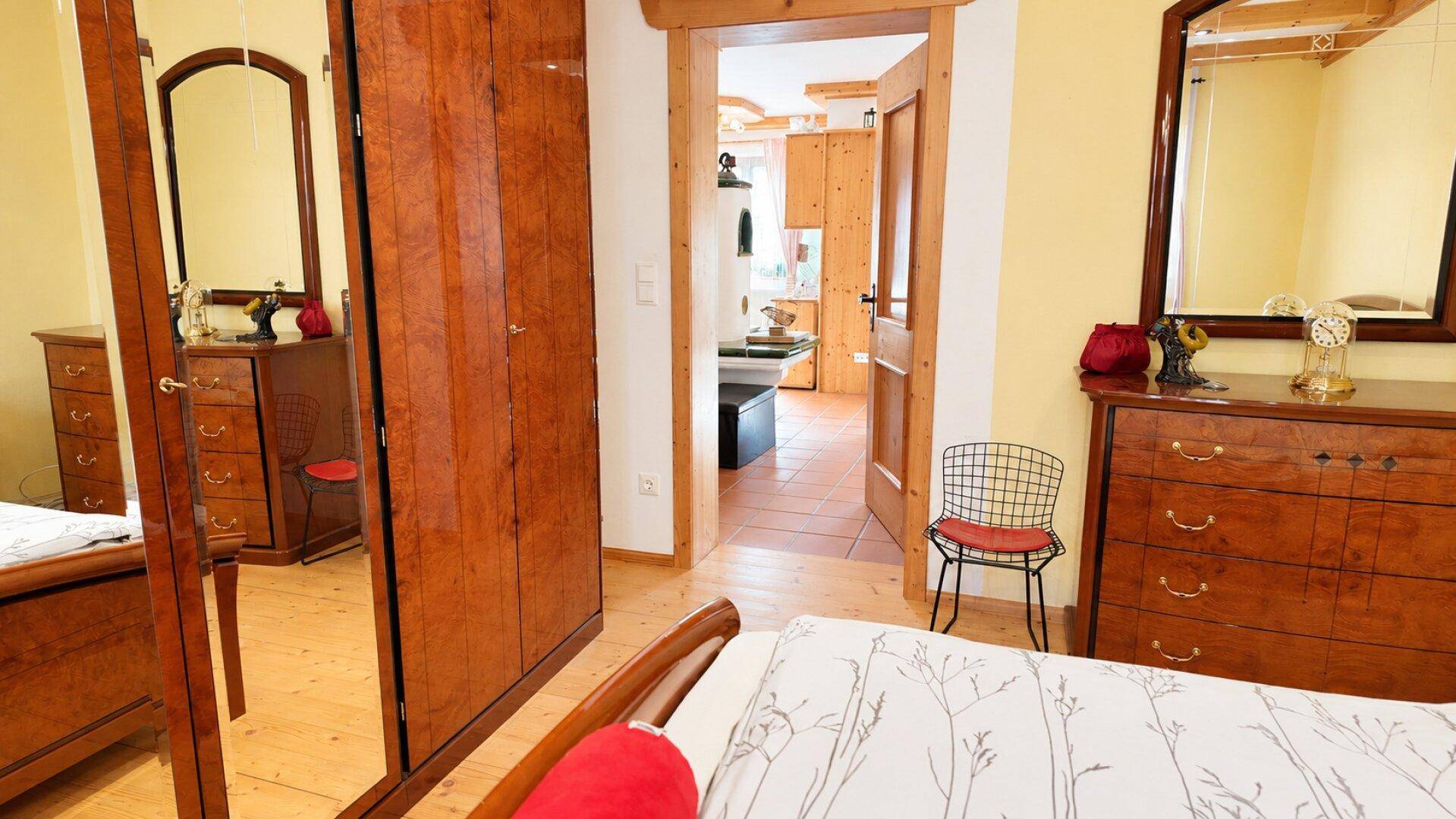 Schlafzimmer mit Holzboden, Spiegelschrank und Blick auf einen angrenzenden Raum mit Kachelofen.