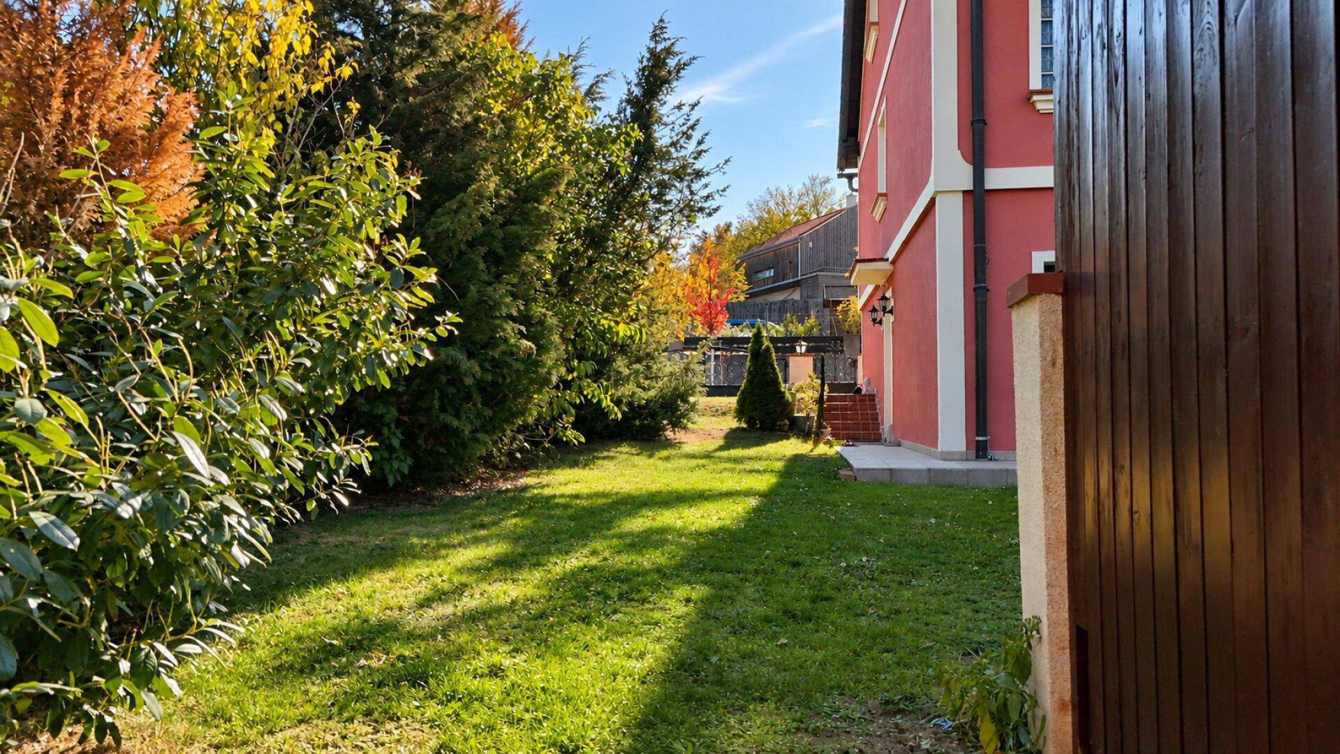 Seitlicher Gartenweg entlang des Hauses mit üppiger Bepflanzung und sonnigem Rasenbereich.