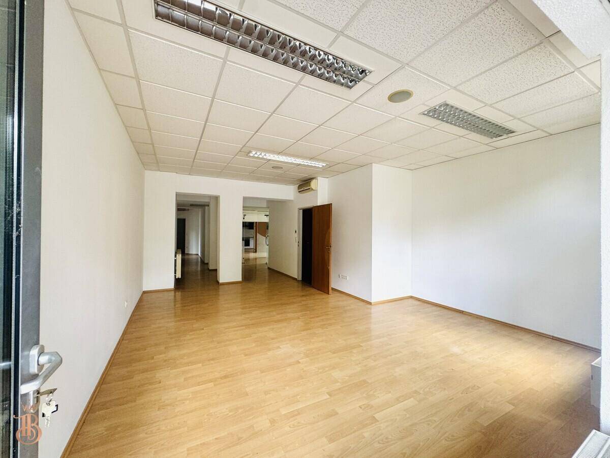 Leerer Büroraum mit Holzfußboden, weißen Wänden und Zugang zu einem Flur.