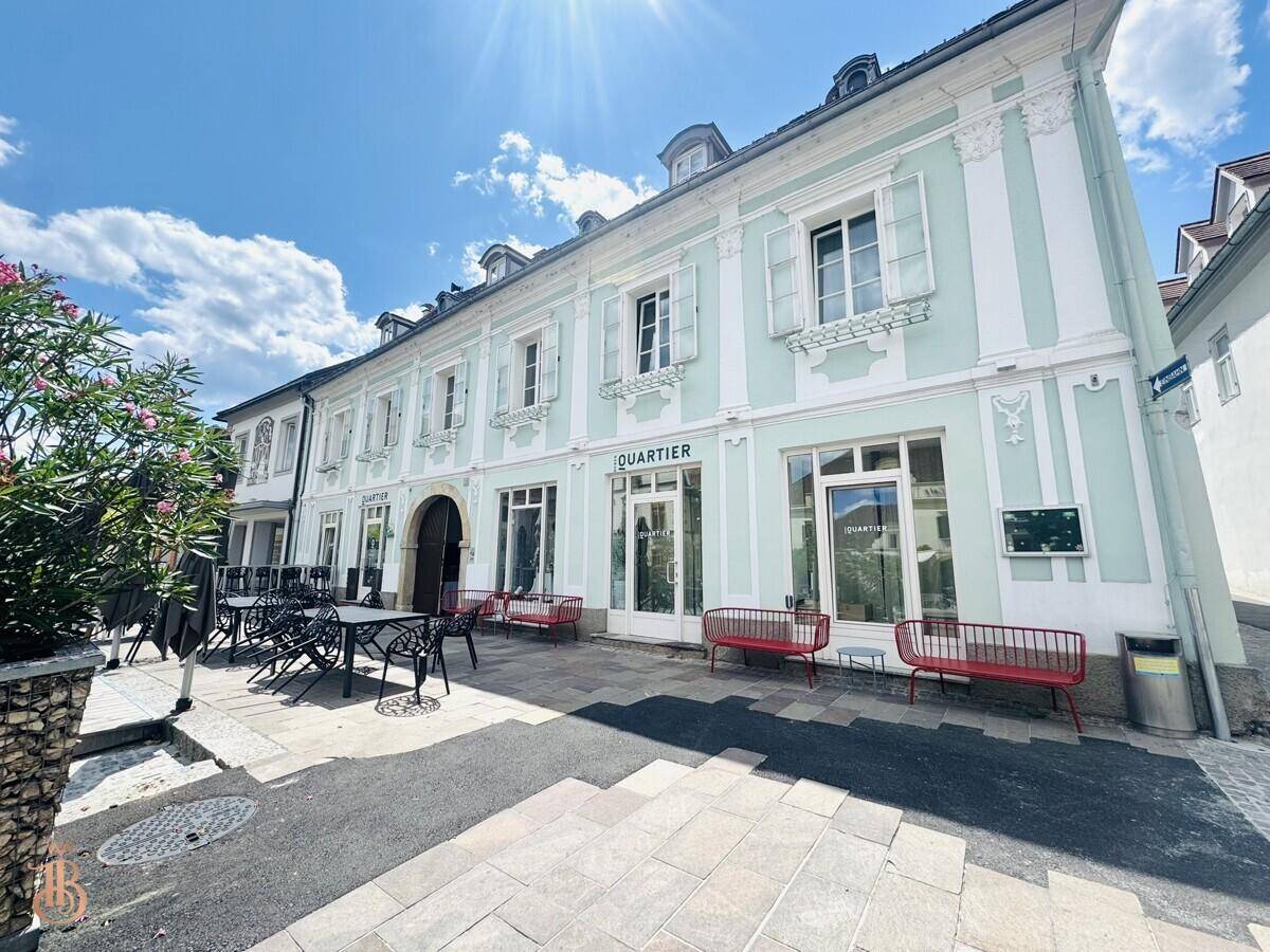 Die Fassade des historischen Gebäudes mit Außenbestuhlung und roten Bänken an einem sonnigen Tag.