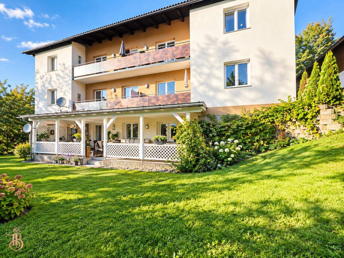 Rückseite des Gebäudes mit mehreren Balkonen und einer überdachten Terrasse, umgeben von Rasenfläche.