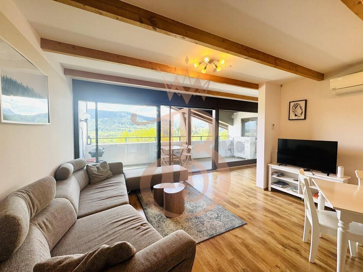 Gemütliches Wohnzimmer mit Sofa, Holzboden und großen Fenstern mit Blick auf die Natur.