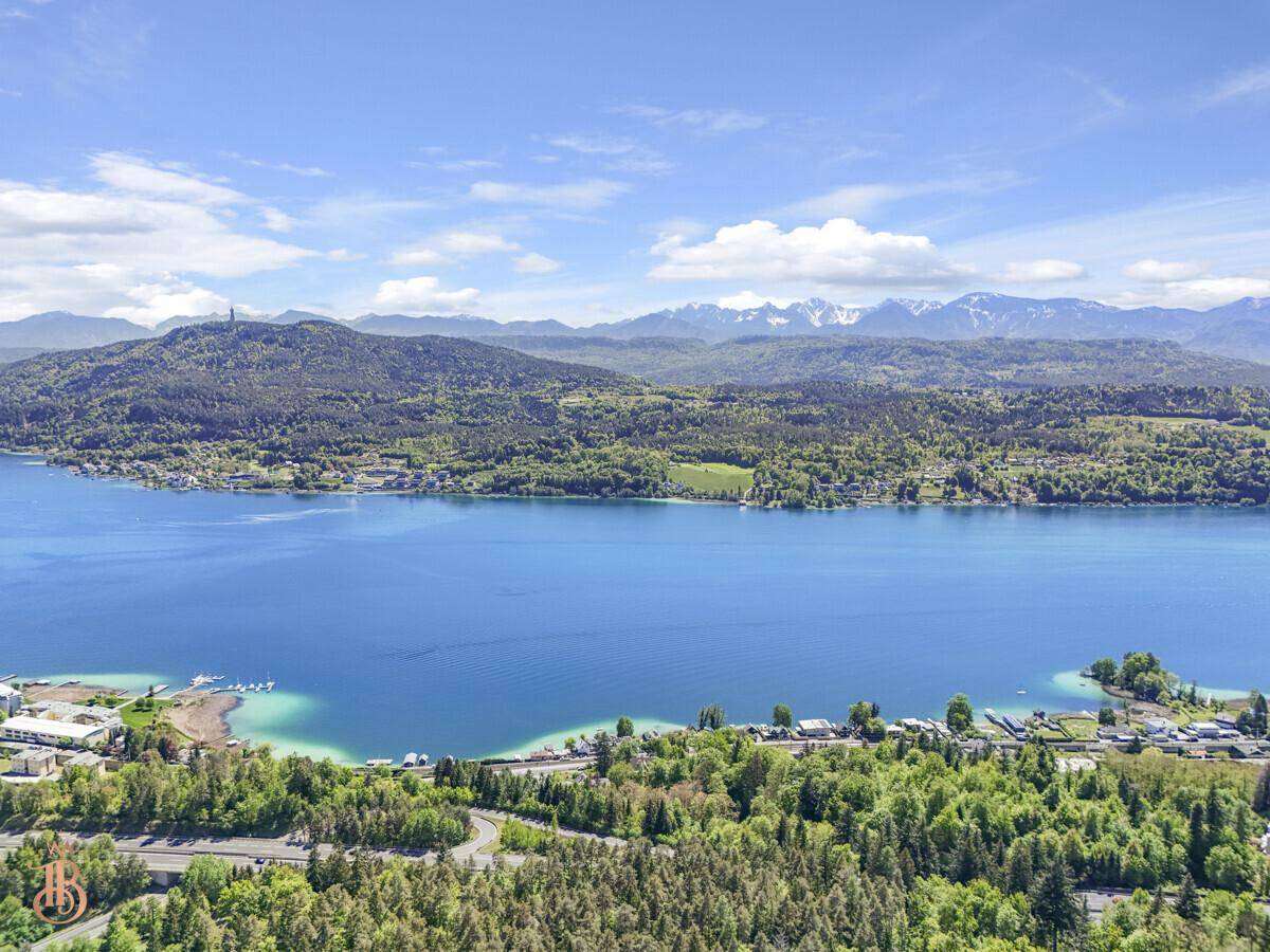 Weitläufiger Luftbildblick auf den tiefblauen See, grüne Ufer und schneebedeckte Berge im Hintergrund.