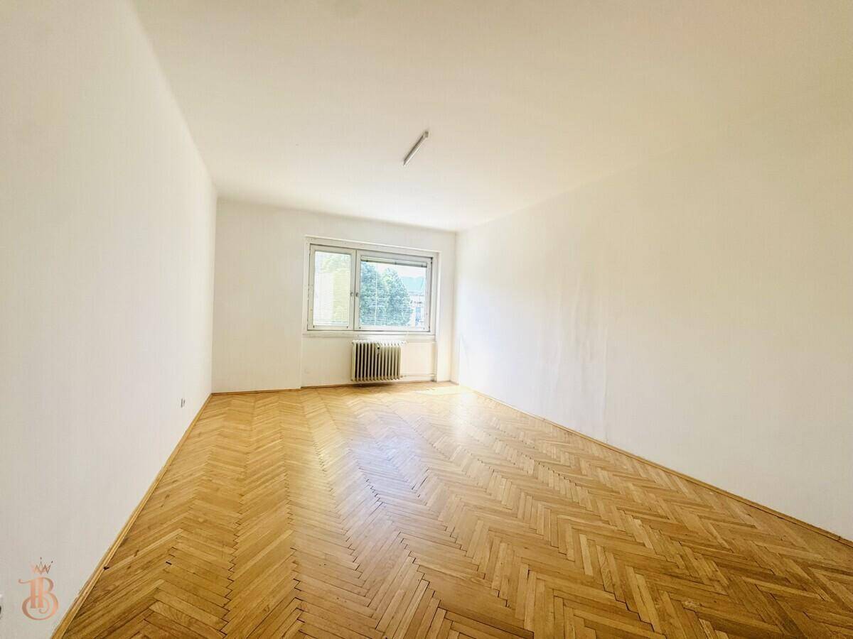 Geräumiges Zimmer mit Fischgrätparkett, weiß gestrichenen Wänden und großem Fenster.