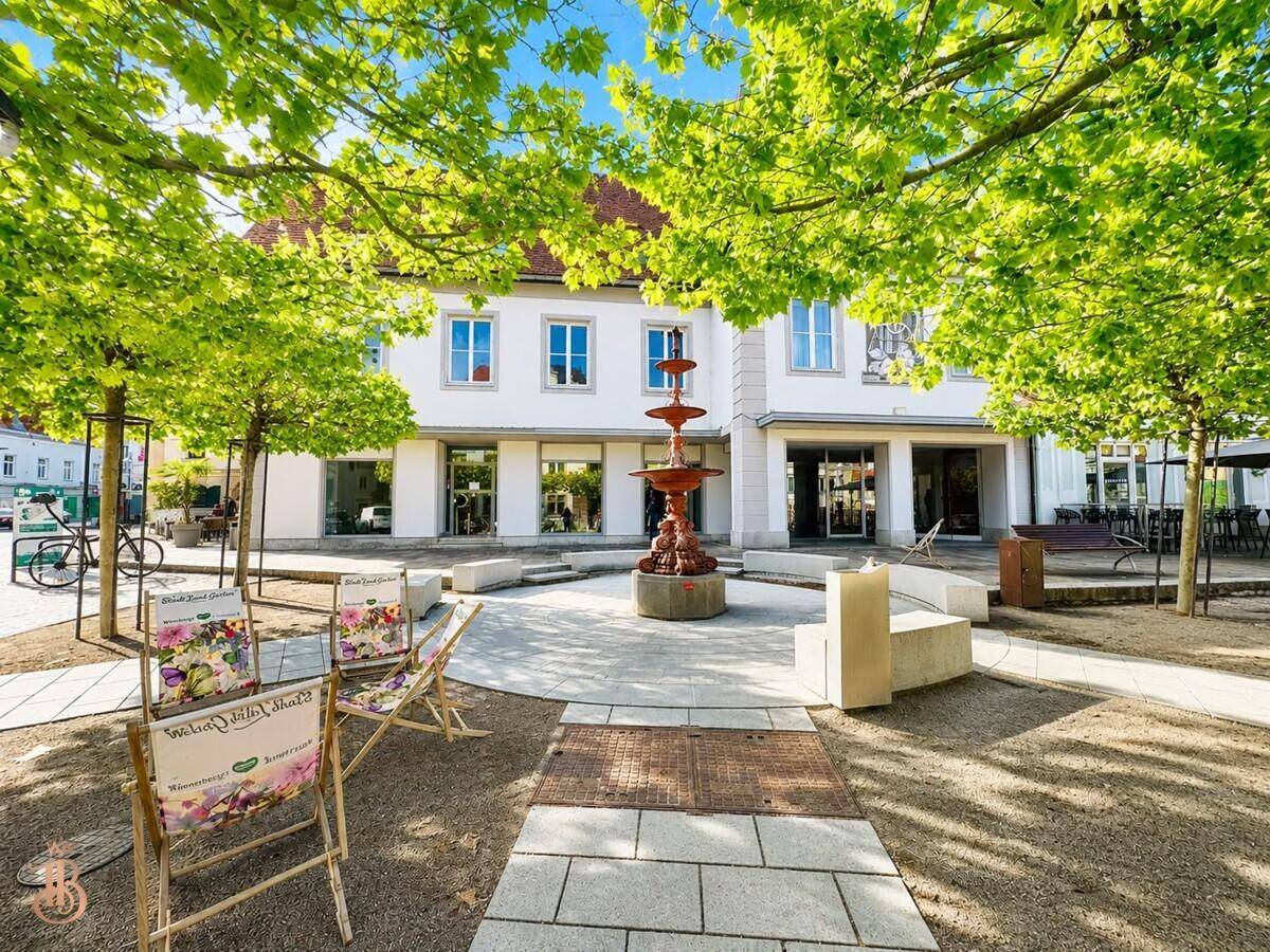 Beleuchteter Stadtplatz mit einem Brunnen und Gebäuden, umgeben von grünen Bäumen.