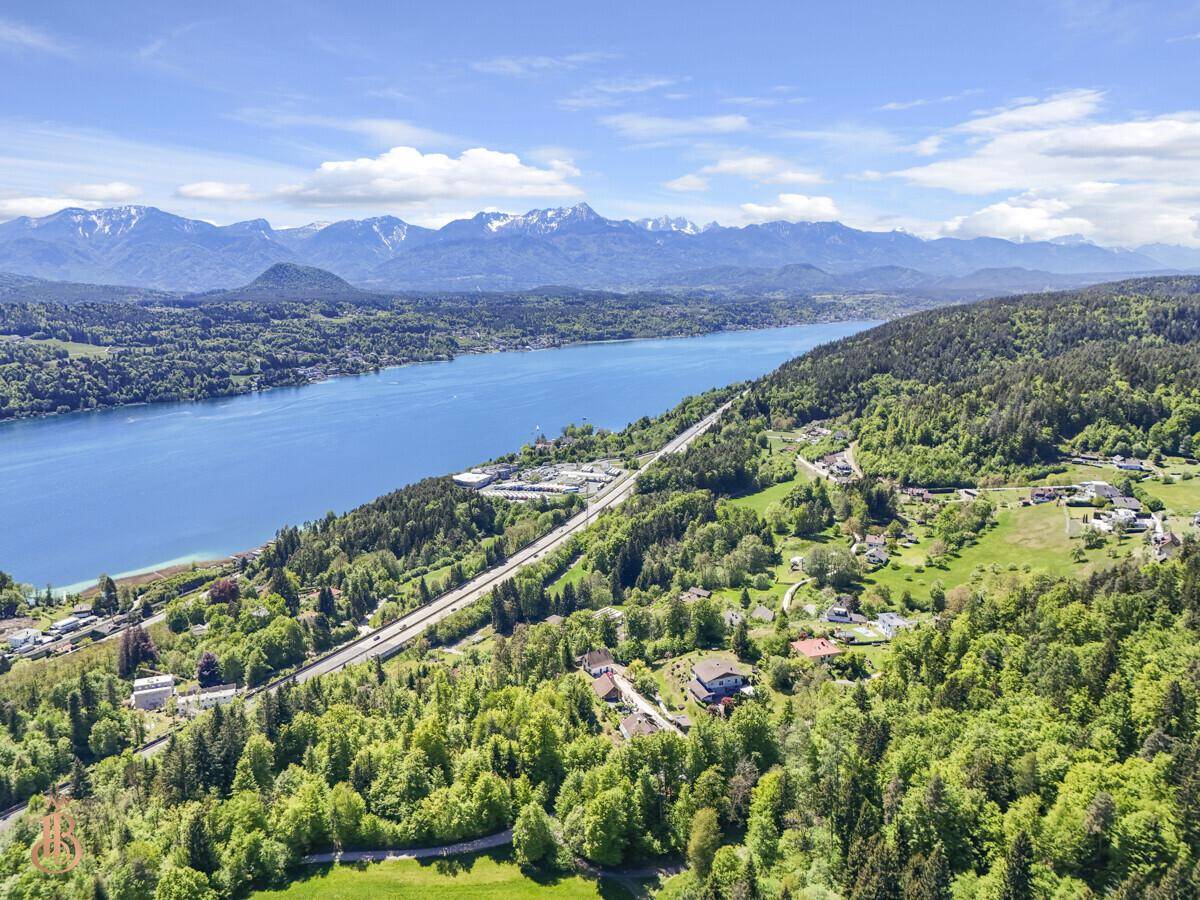 Ausgedehnte Luftaufnahme des Sees mit Uferstraße, bewaldeten Hängen und weitläufiger Landschaft.