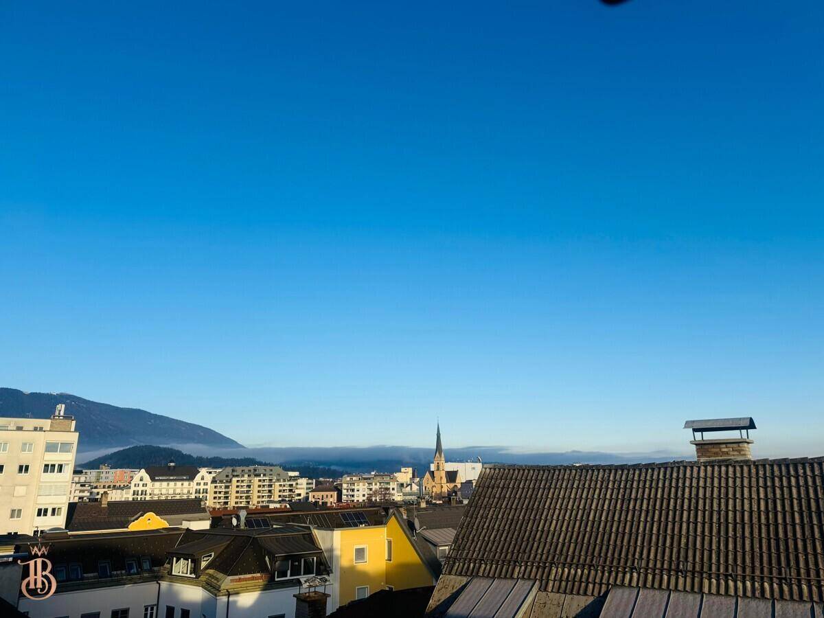 Ausblick über die städtische Bebauung mit einem Kirchturm und Bergen in der Ferne.