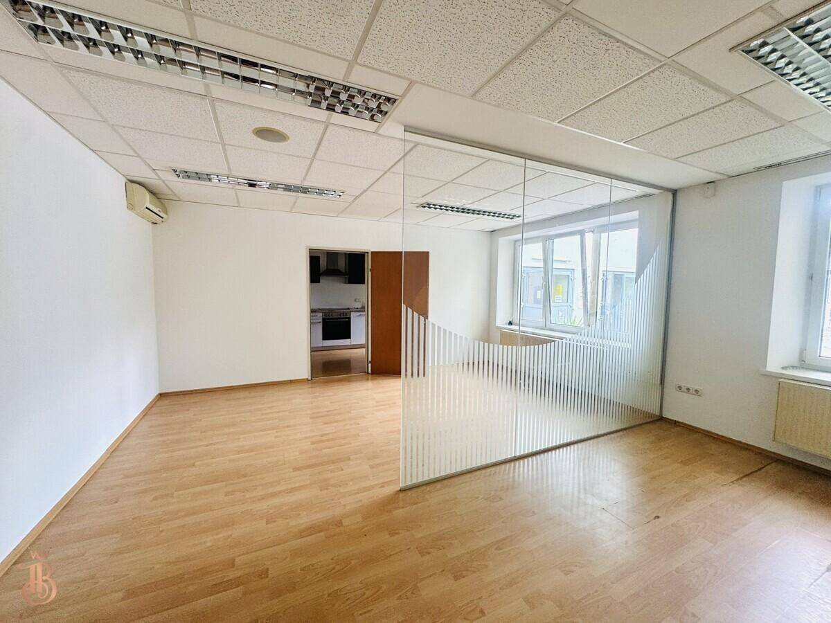 Büroraum mit Holzfußboden, Glasabtrennung und Blick auf eine kleine Küchenzeile.