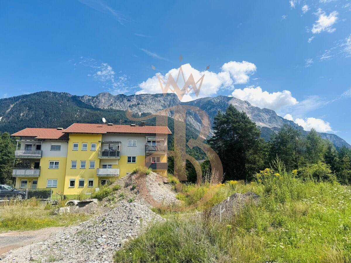 Mehrfamilienhaus mit Balkonen und gelber Fassade, umgeben von Bergen und grüner Landschaft.