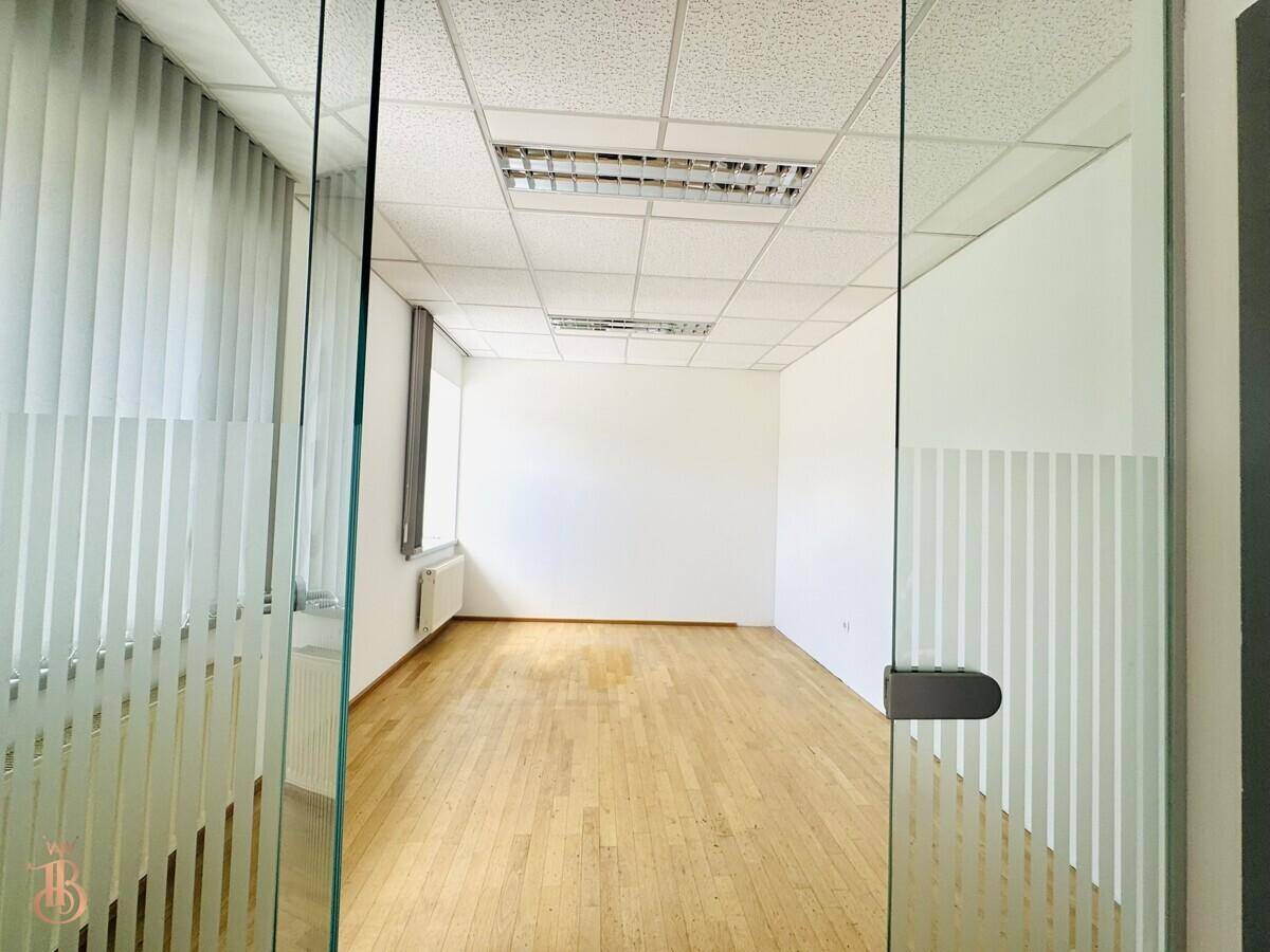 Blick durch die Glastür in einen leeren Büroraum mit Holzfußboden und Fenstern.