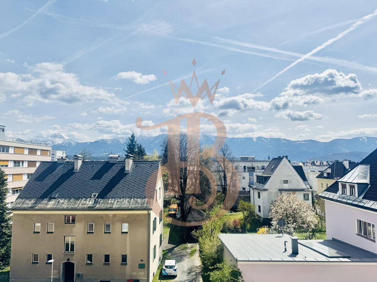 Blick auf eine ruhige Wohnstraße mit Häusern, Bäumen und schneebedeckten Bergen im Hintergrund.