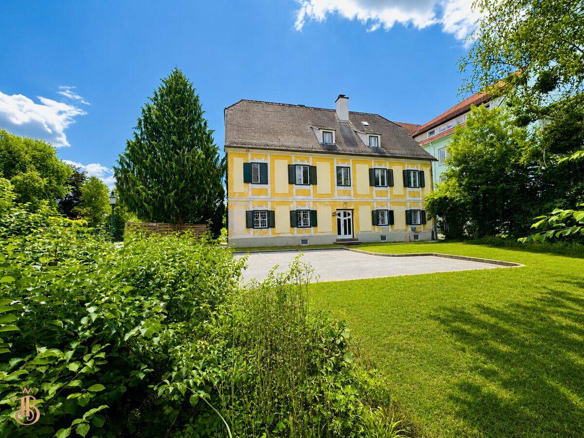 Gelbes historisches Gebäude mit grünen Fensterläden und weitläufigem Vorgarten unter blauem Himmel.
