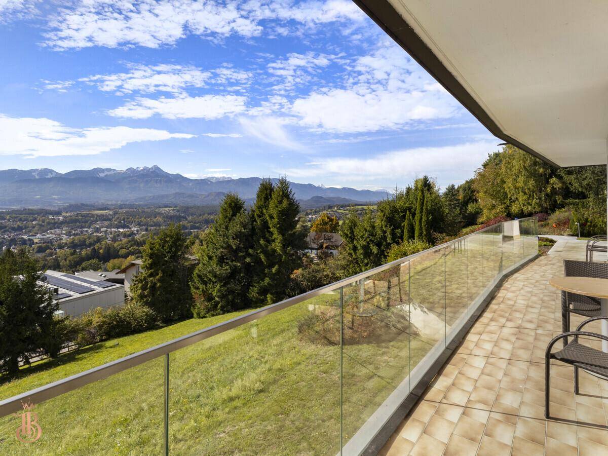 Sonniger Balkon mit Sitzgelegenheiten und weitem Blick über die grüne Landschaft zum See.