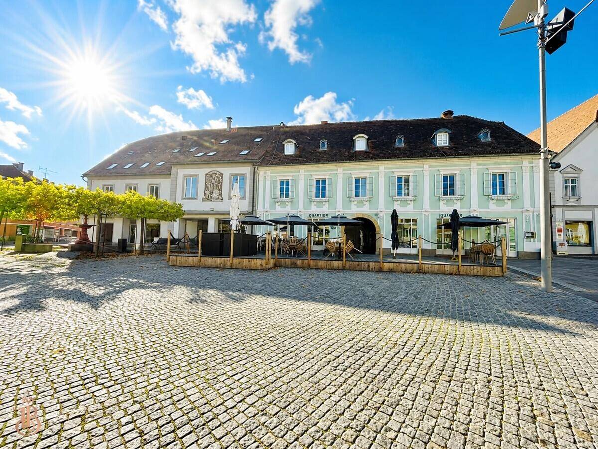 Sonniger Stadtplatz mit Kopfsteinpflaster und historischen Gebäuden, darunter ein Restaurant mit Außenterrasse.
