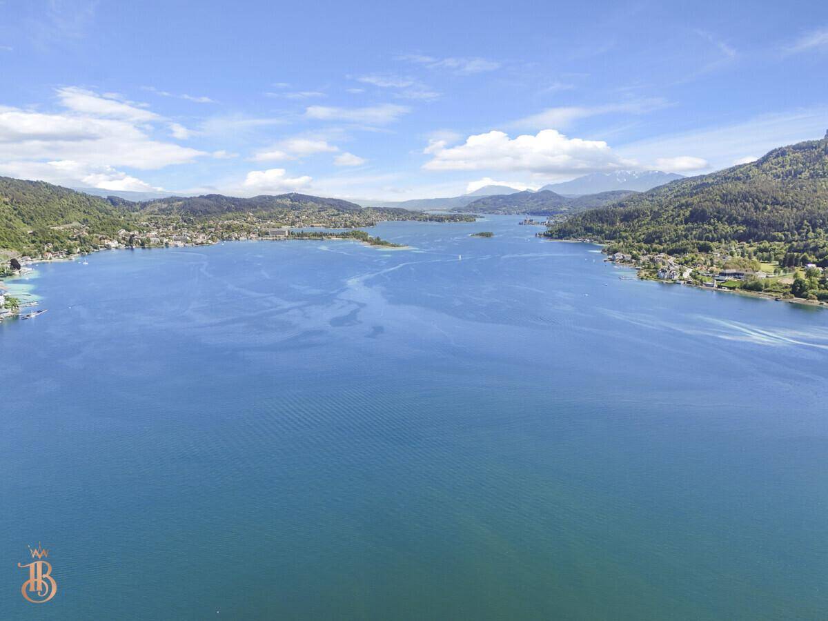 Panoramische Luftaufnahme des langgestreckten Sees, umgeben von grünen Hügeln und fernen Bergen.