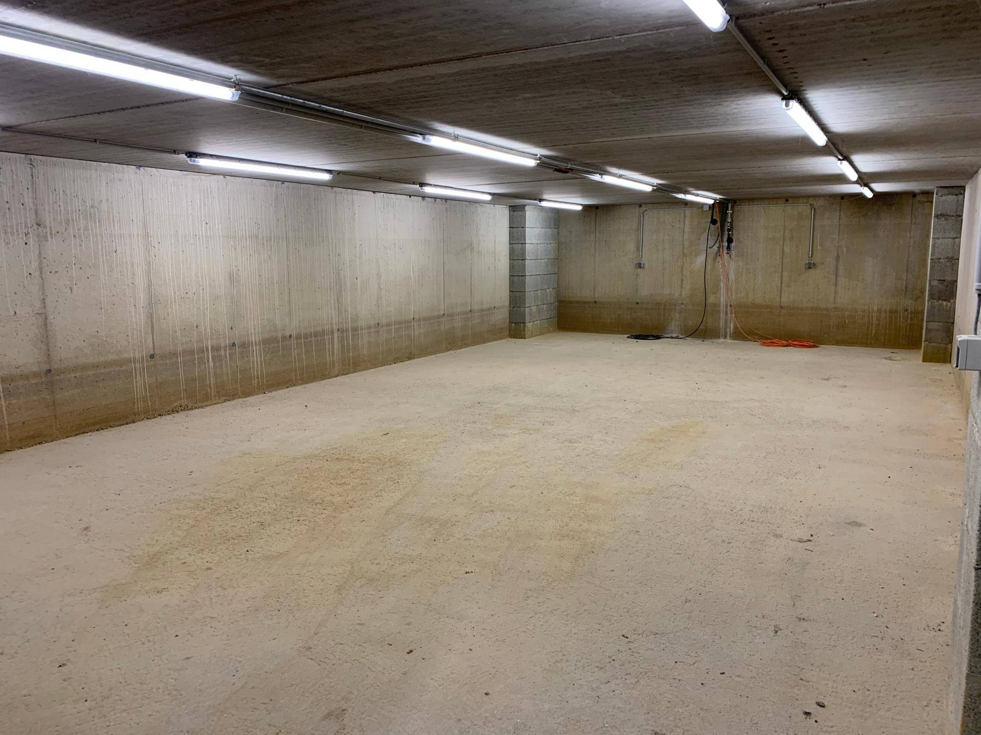 Langer, leerer Kellerraum mit Betonwänden und -boden, ausgestattet mit Deckenbeleuchtung.