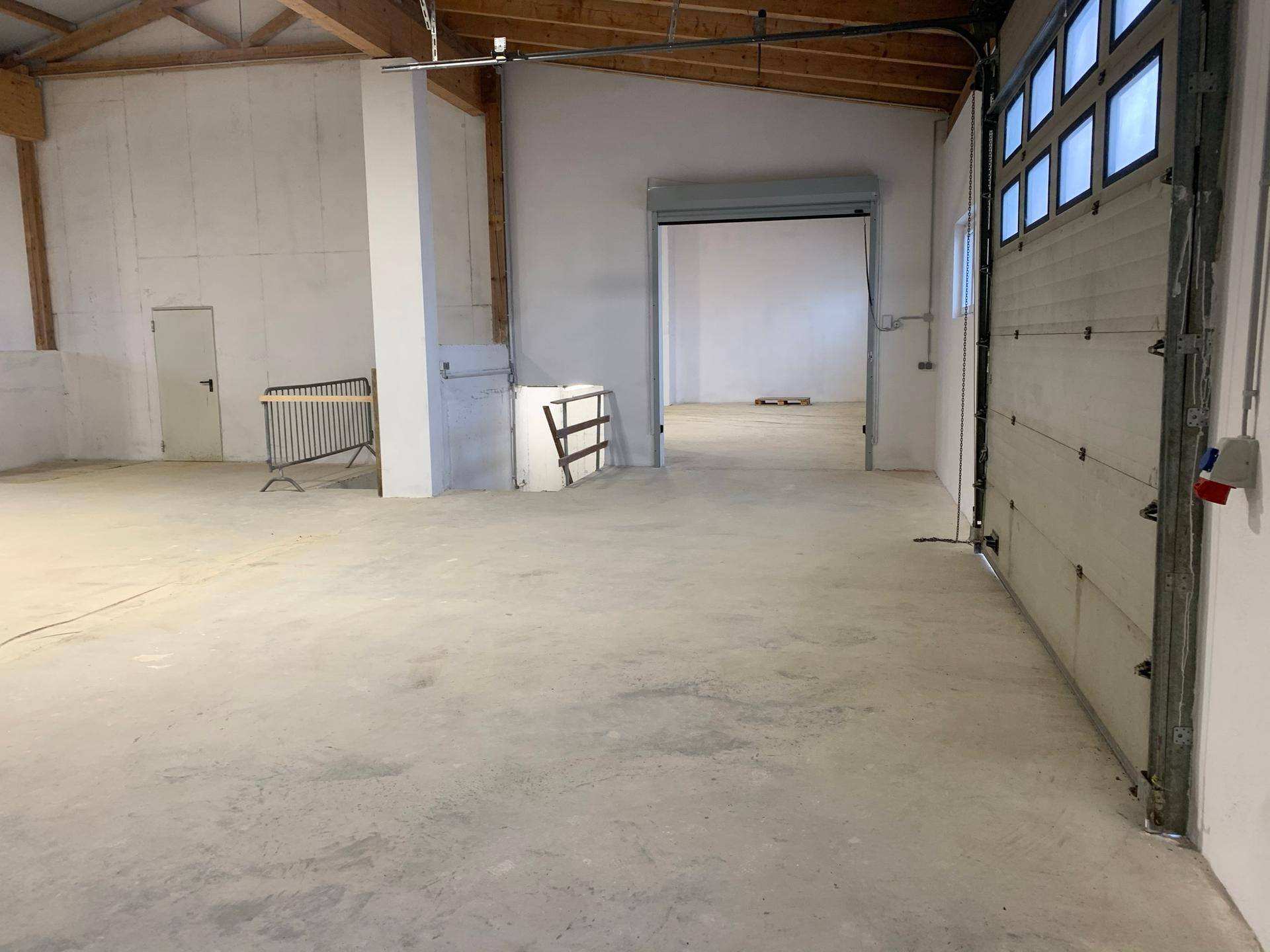 Innenansicht einer Lagerhalle mit Rolltor, Betonboden und Zugang zu weiteren Bereichen.