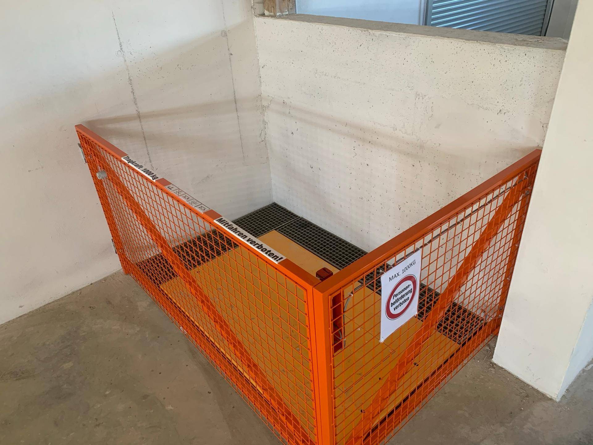 Lastenaufzug in einem Kellerbereich mit orangefarbenem Schutzgitter und Warnhinweisen.