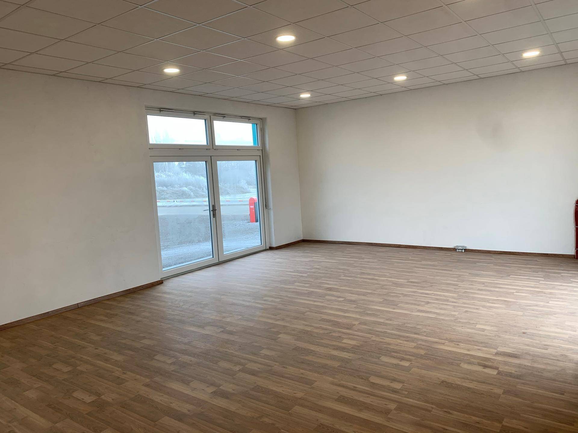 Heller, leerer Raum mit Laminatboden, großen Fenstern und Einbaustrahlern in der Decke.