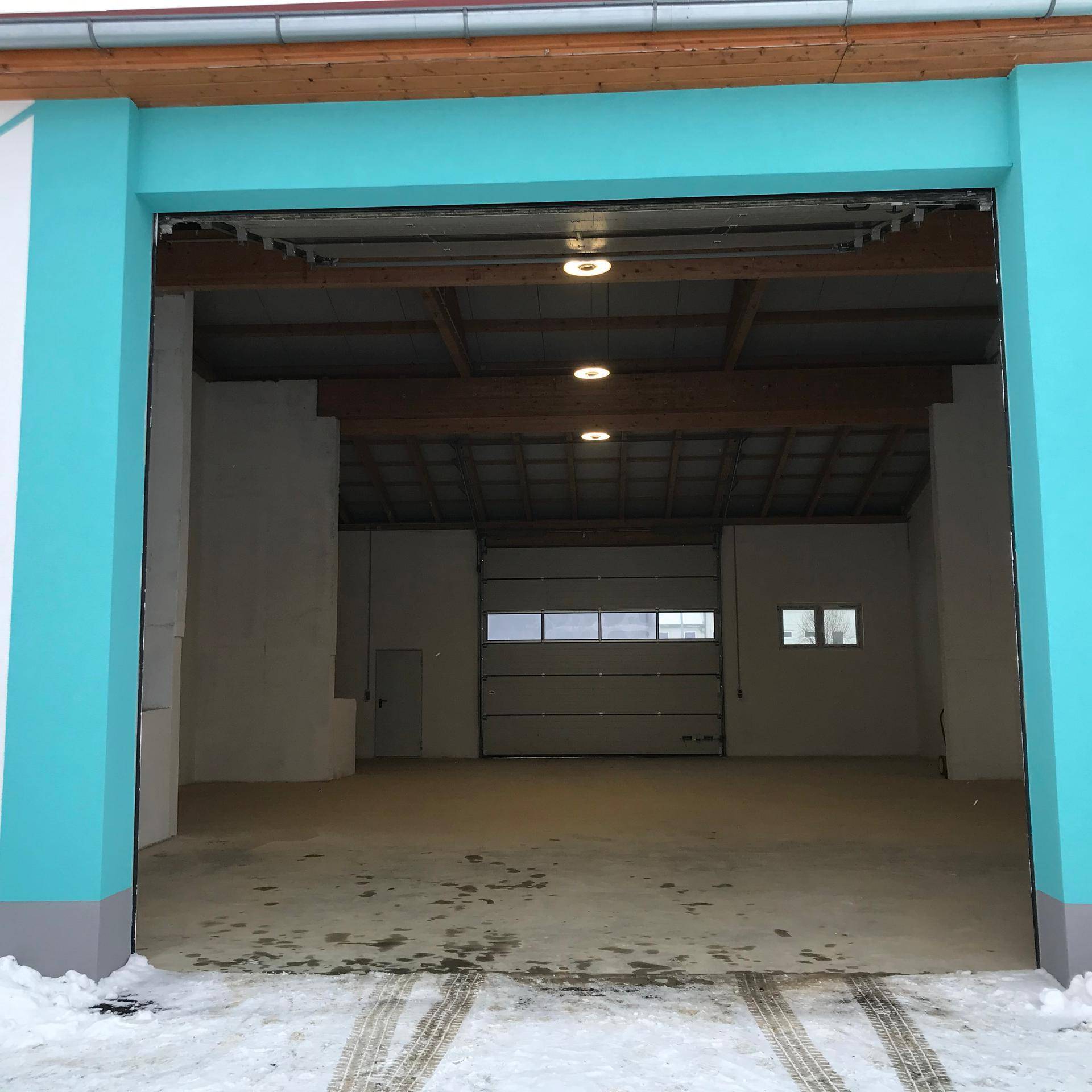 Eingangsbereich einer Halle mit Rolltor und türkisfarbener Fassade im Winter.