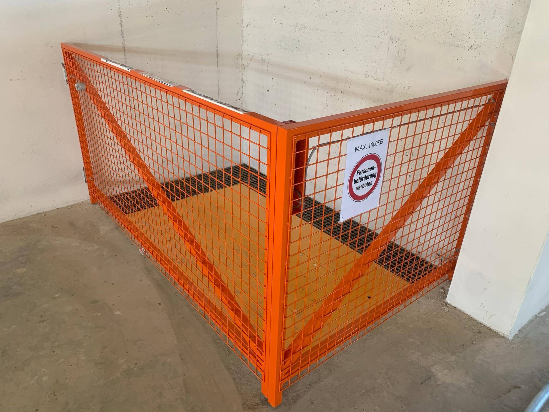 Lastenaufzug in einem Kellerbereich mit orangefarbenem Schutzgitter und Warnhinweisen.