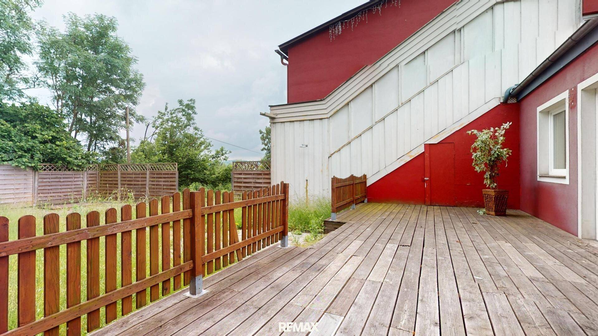 Geräumige Holzterrasse mit rustikalem Holzzaun, angrenzend an das rote Gebäude und eine Grünfläche.