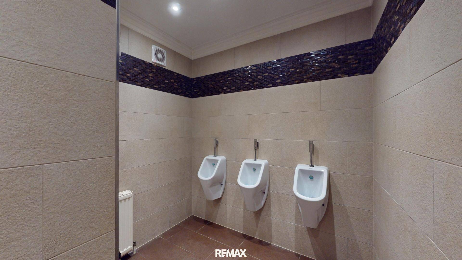 Öffentlicher Toilettenraum mit drei Urinalen, beigefarbenen Fliesen und einer dekorativen Mosaikbordüre.