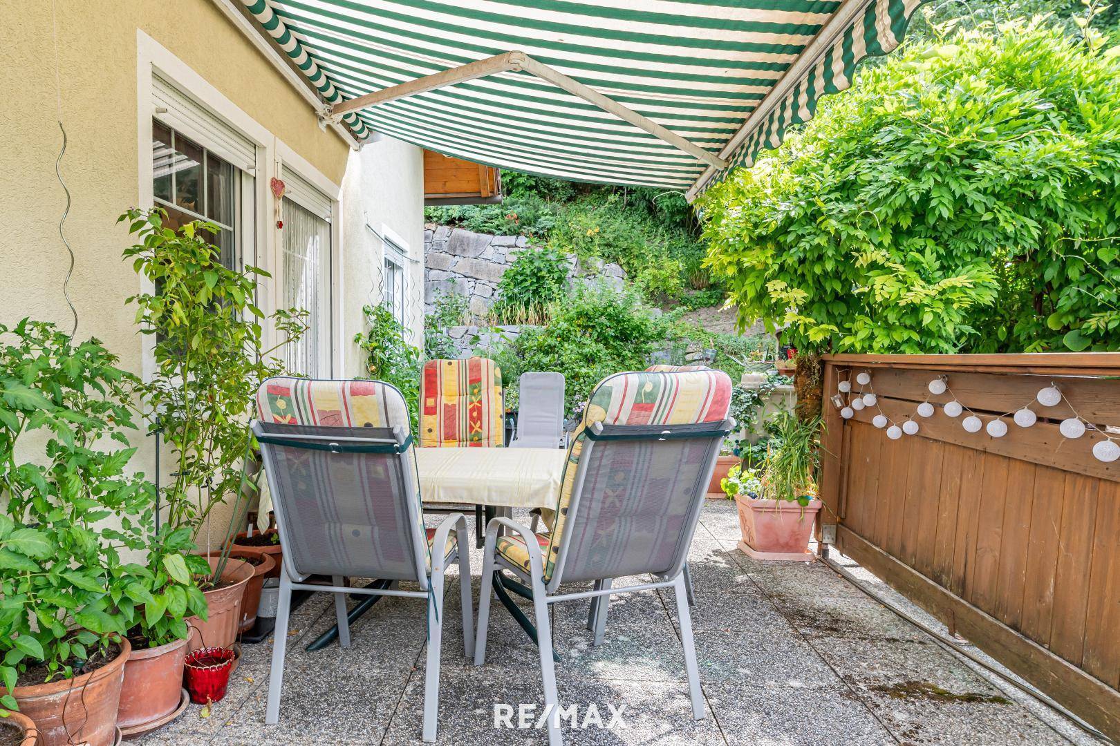 Gemütliche Terrasse mit Markise und Gartenmöbeln, umgeben von üppigem Grün und Topfpflanzen.